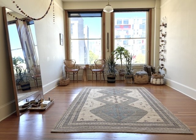 Earth Studio is a bright, welcoming room with large windows, potted plants, woven baskets, wooden chairs, a patterned rug, and a full-length mirror. Ample yoga mats, blankets, and blocks are provided, and folding chairs for group therapy or events.