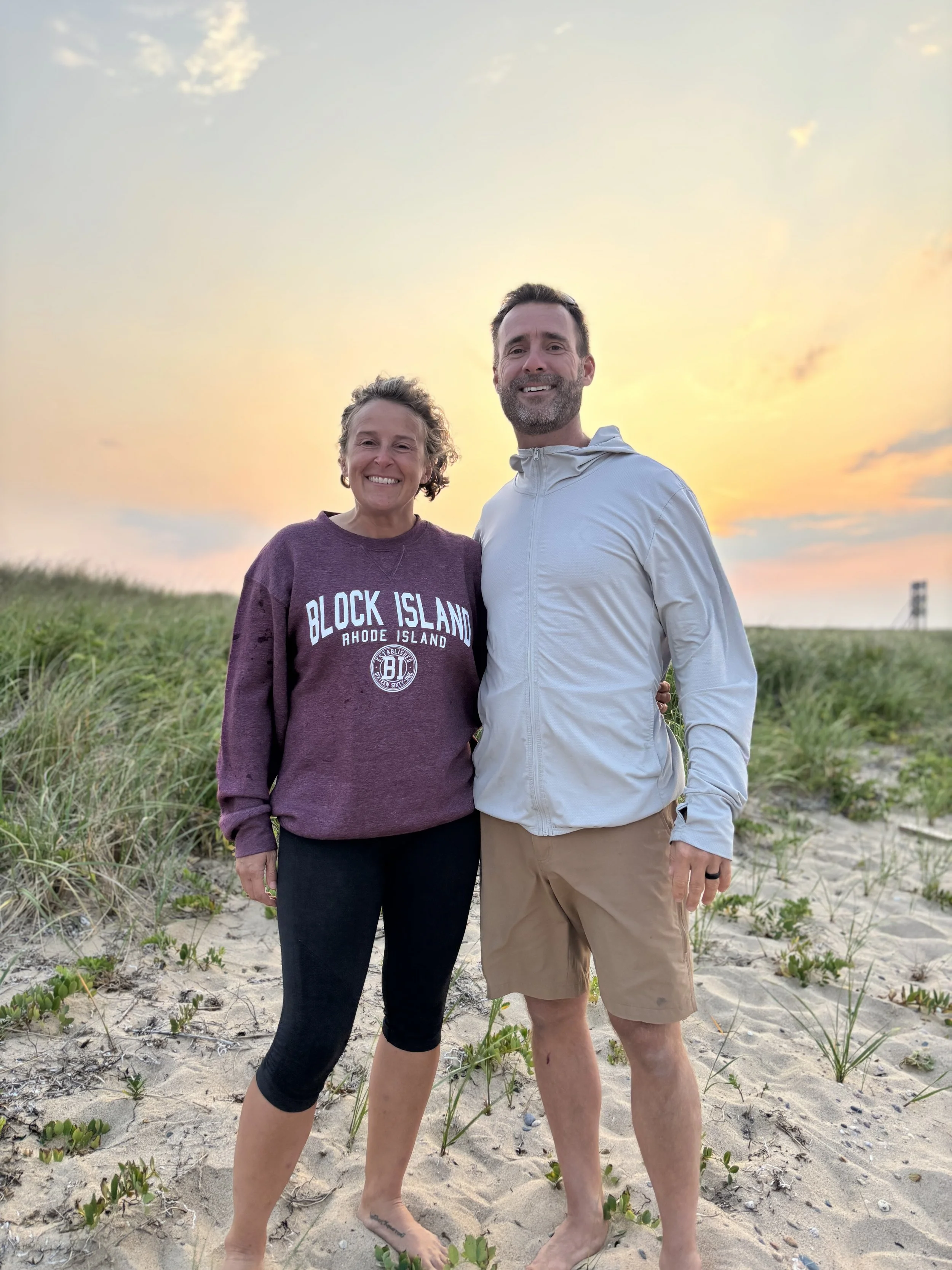 A smiling couple standing on a sandy beach with grass and dunes, during sunset, with a sky of pastel pink and orange colors.