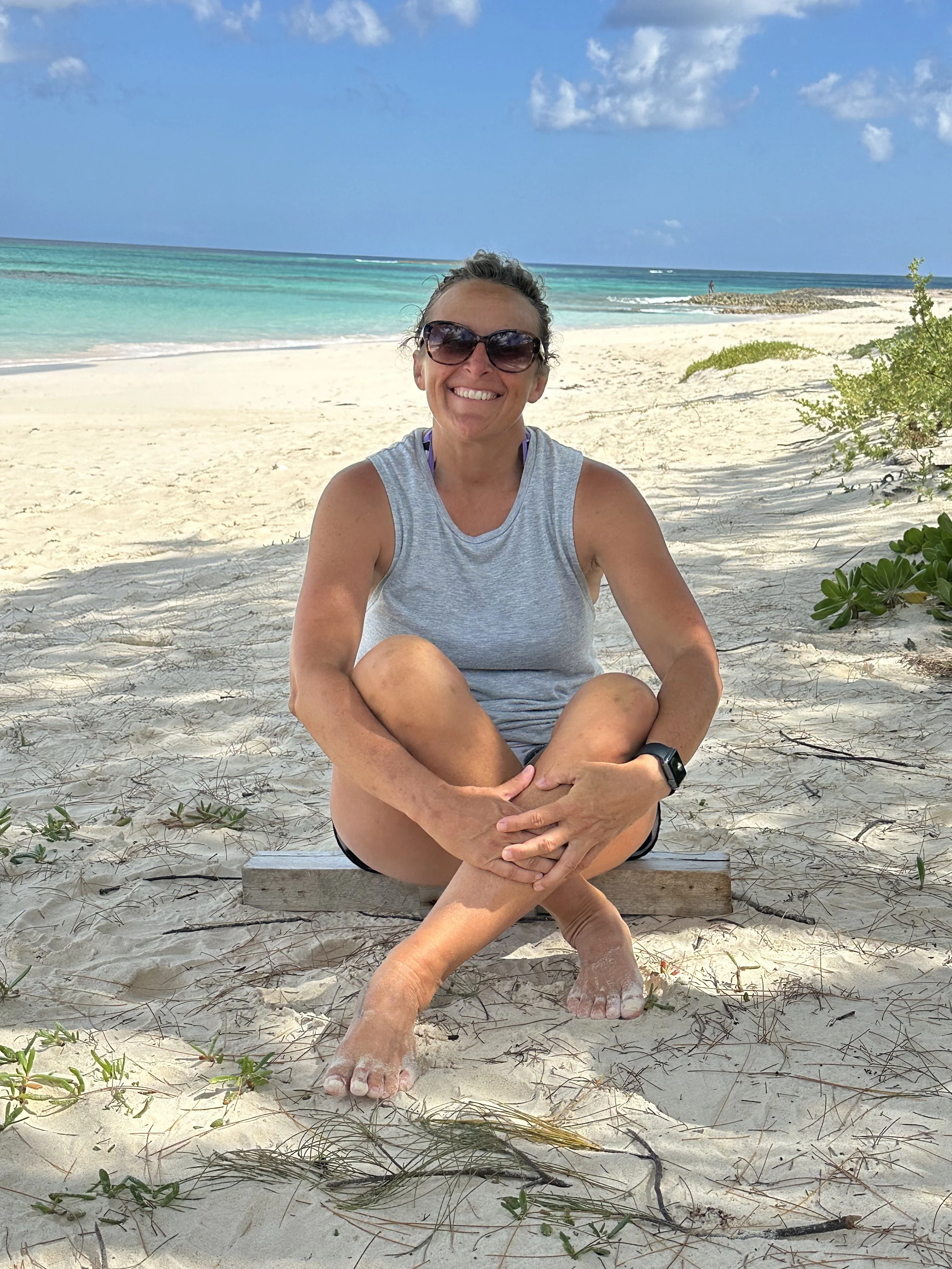 A woman sitting on the sandy beach with the ocean and blue sky behind her, wearing sunglasses and a gray sleeveless top, smiling.