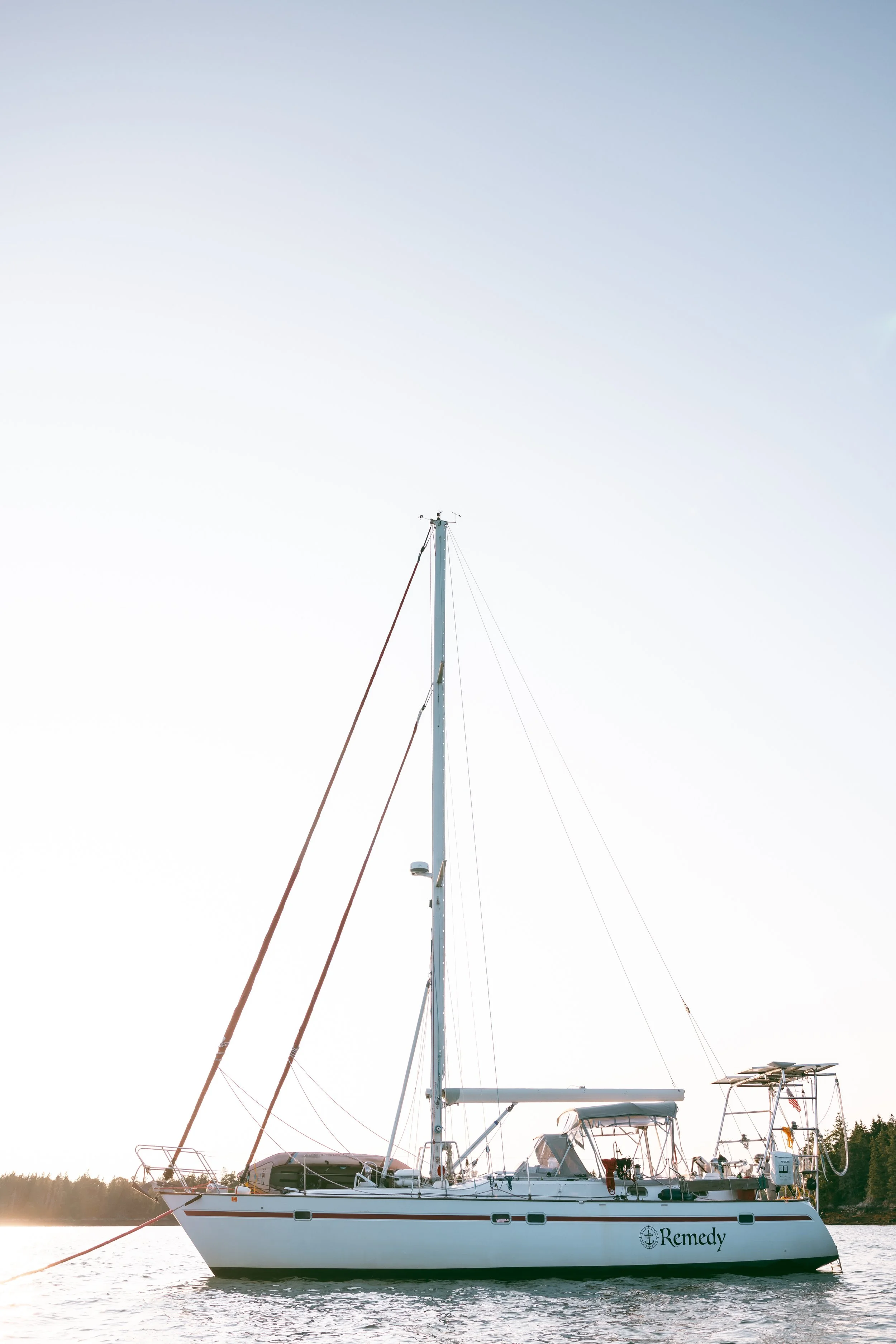 Sailboat named Remedy floating on water near forested shoreline at sunset