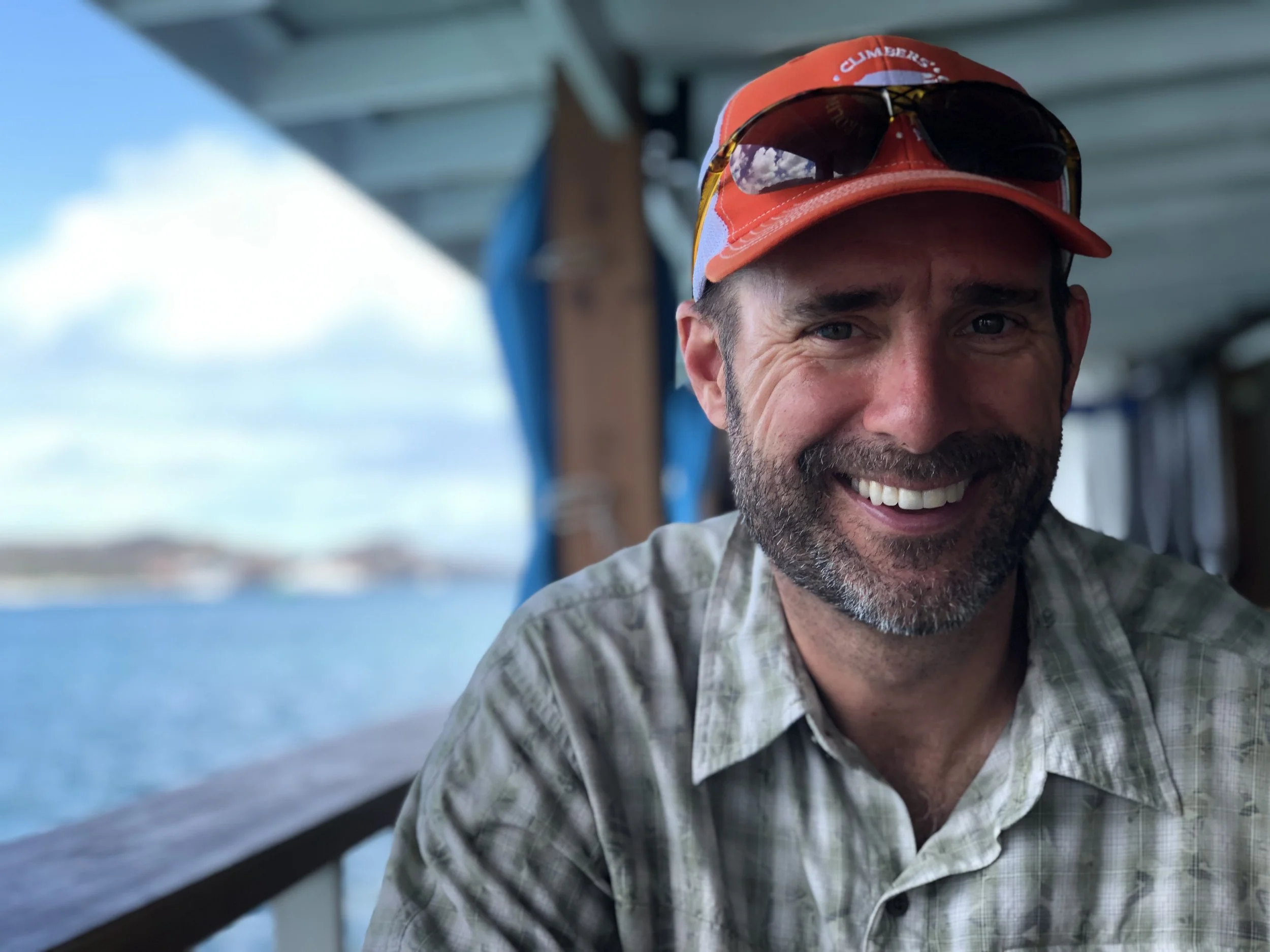 Smiling man with sunglasses on his red cap, wearing a plaid shirt, sitting outdoors near water under a shelter.