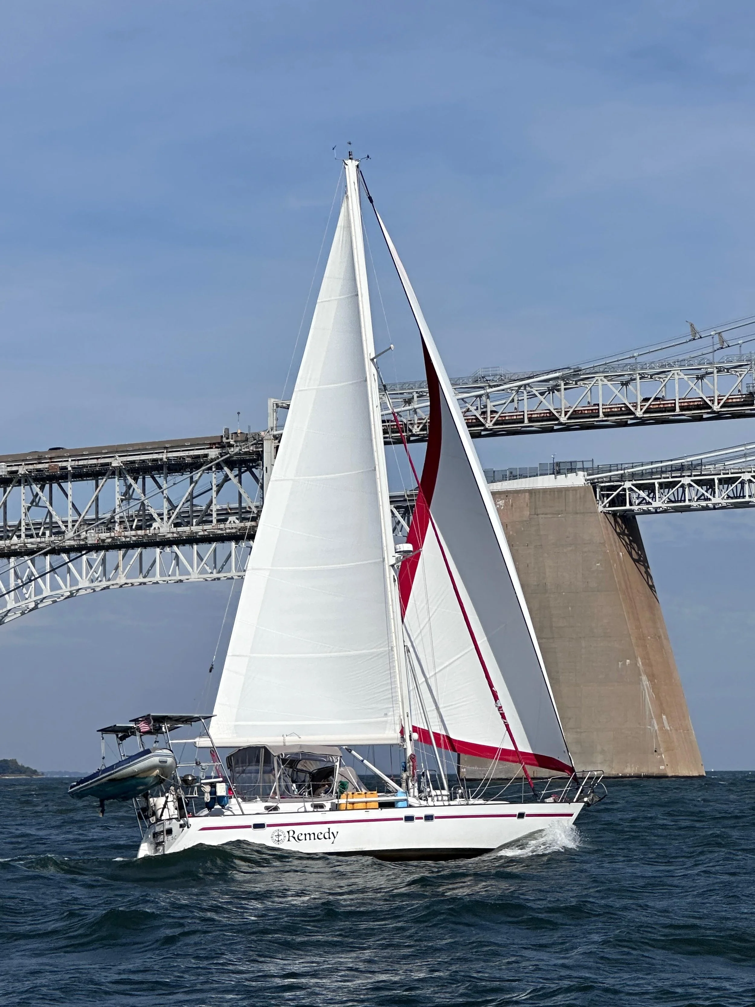 Sailing boat named Remedy on water with a large bridge in background under clear blue sky.