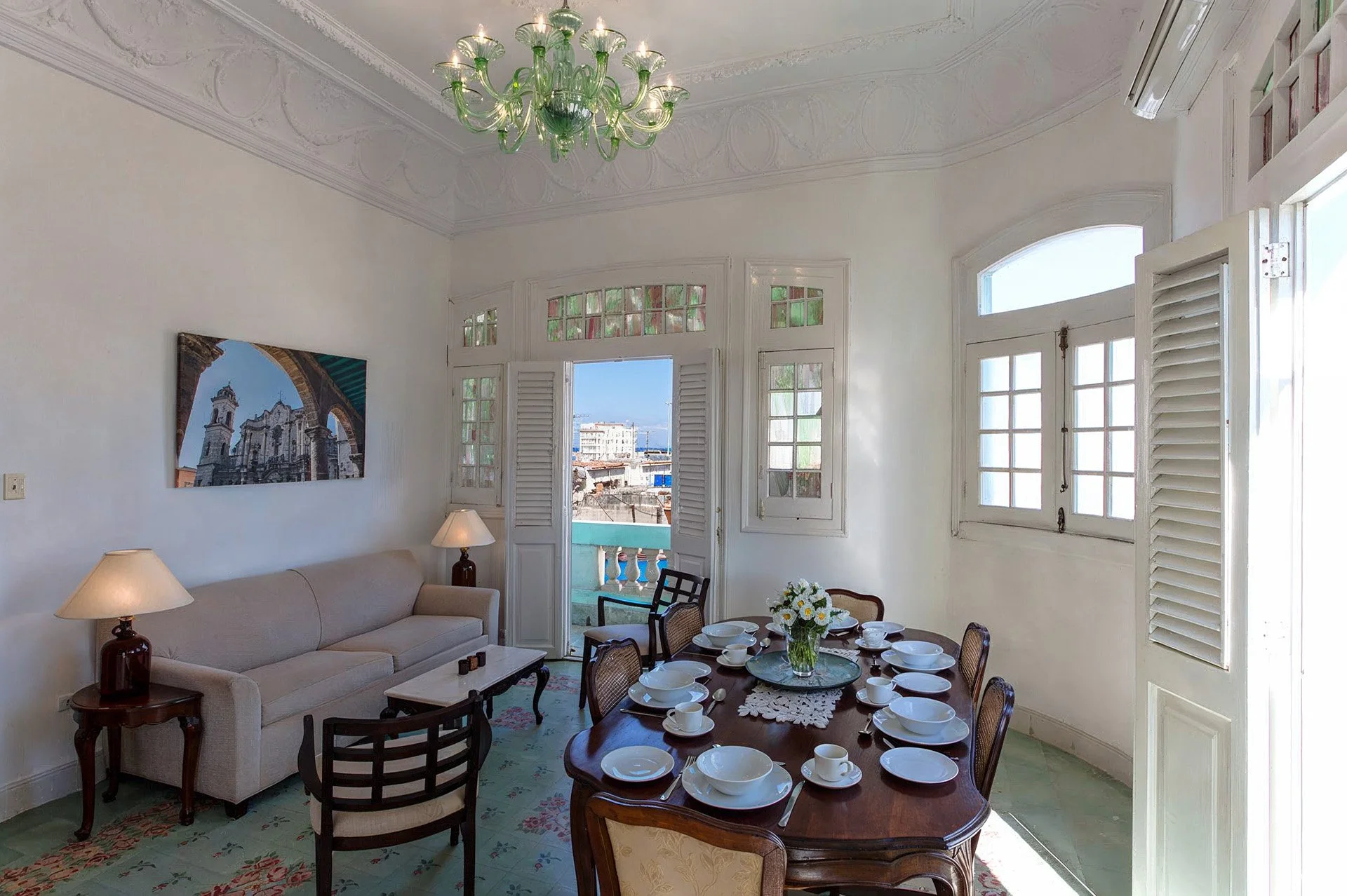 Cuba, Havana - Casa Concordia Room with a dining table set for breakfast, a beige couch with lamps on side tables, and a balcony with city view through open French doors and windows, bright sunlight, chandelier, white walls, decorative crown molding.