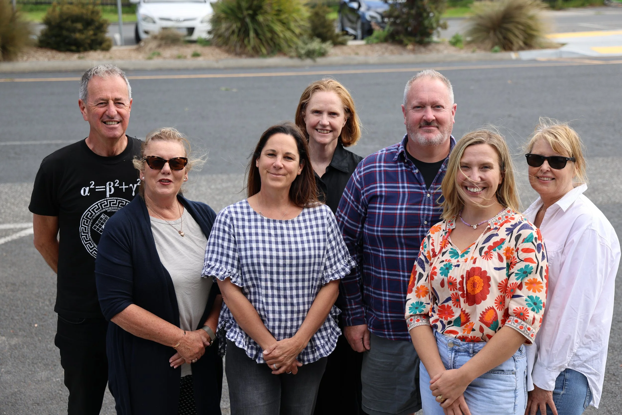 Group of seven people standing outdoors on pavement, smiling at the camera, with parked cars and bushes in the background.