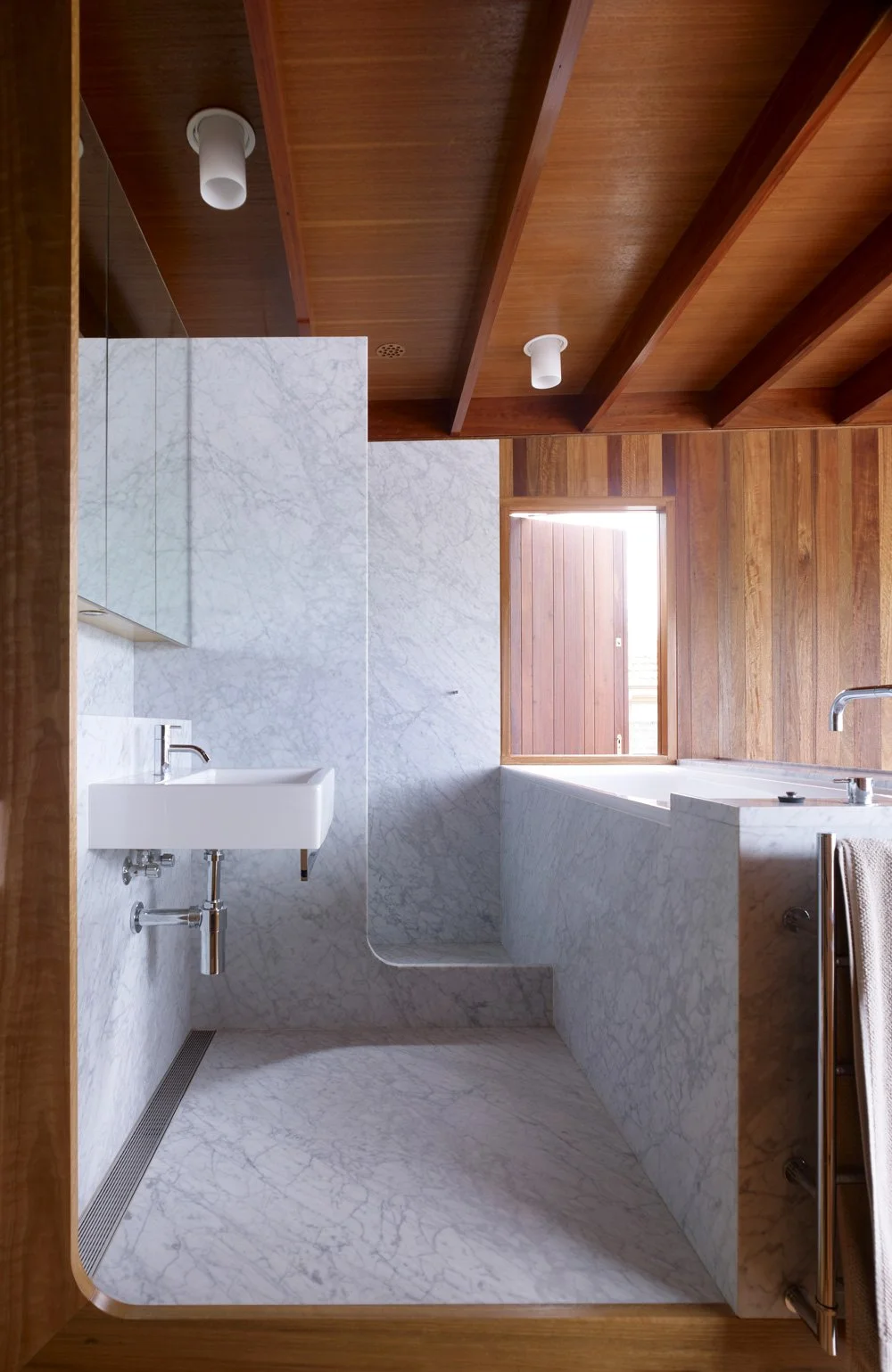 A modern bathroom with marble walls and floor, wooden ceiling with exposed beams, a rectangular window, and minimal fixtures including a small sink, faucet, and towel rack.