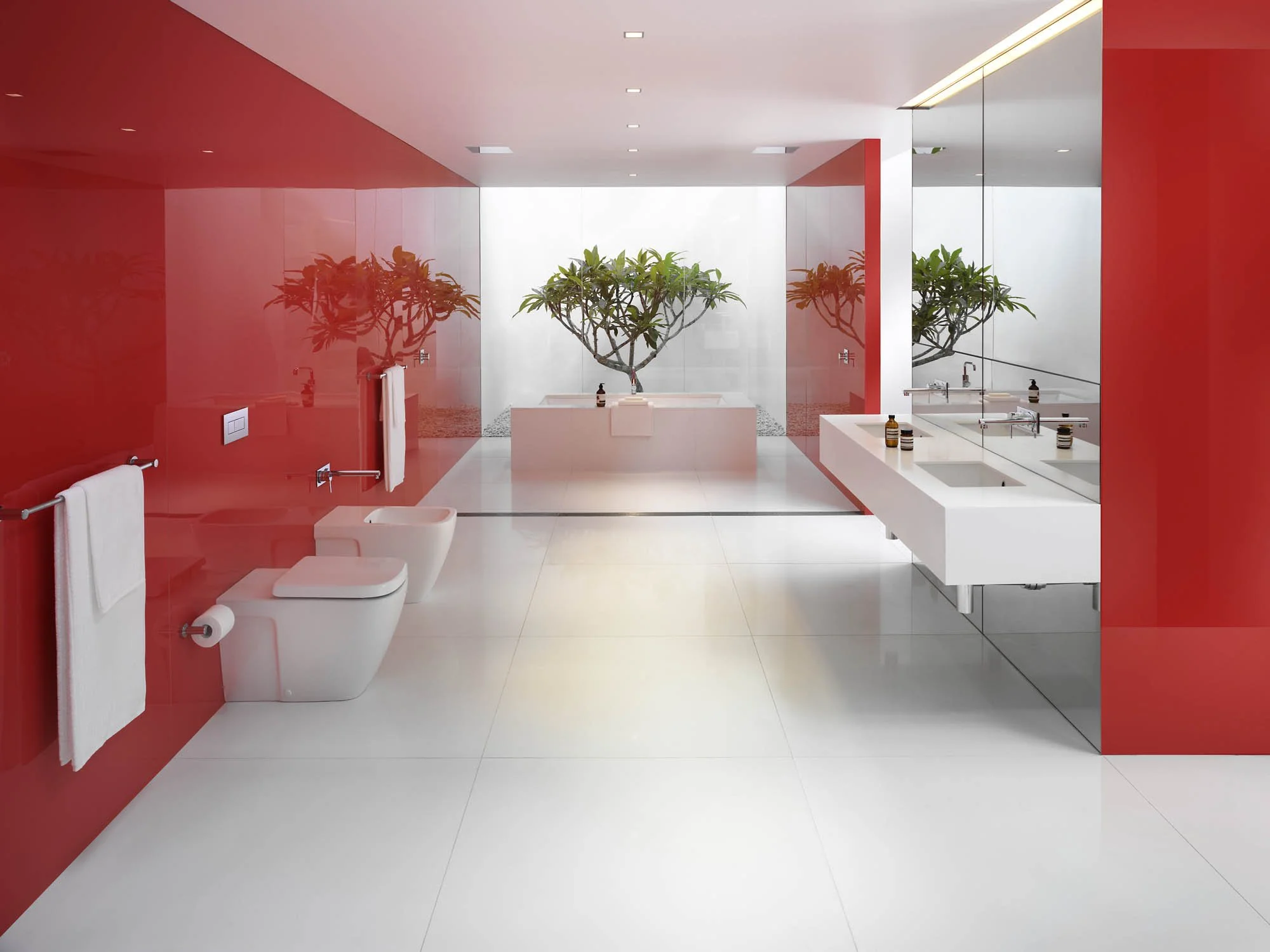 Modern bathroom with white flooring and red walls, featuring a bidet and toilet on the left, a double sink with bottles on the right, and a bathtub with indoor plants at the back.