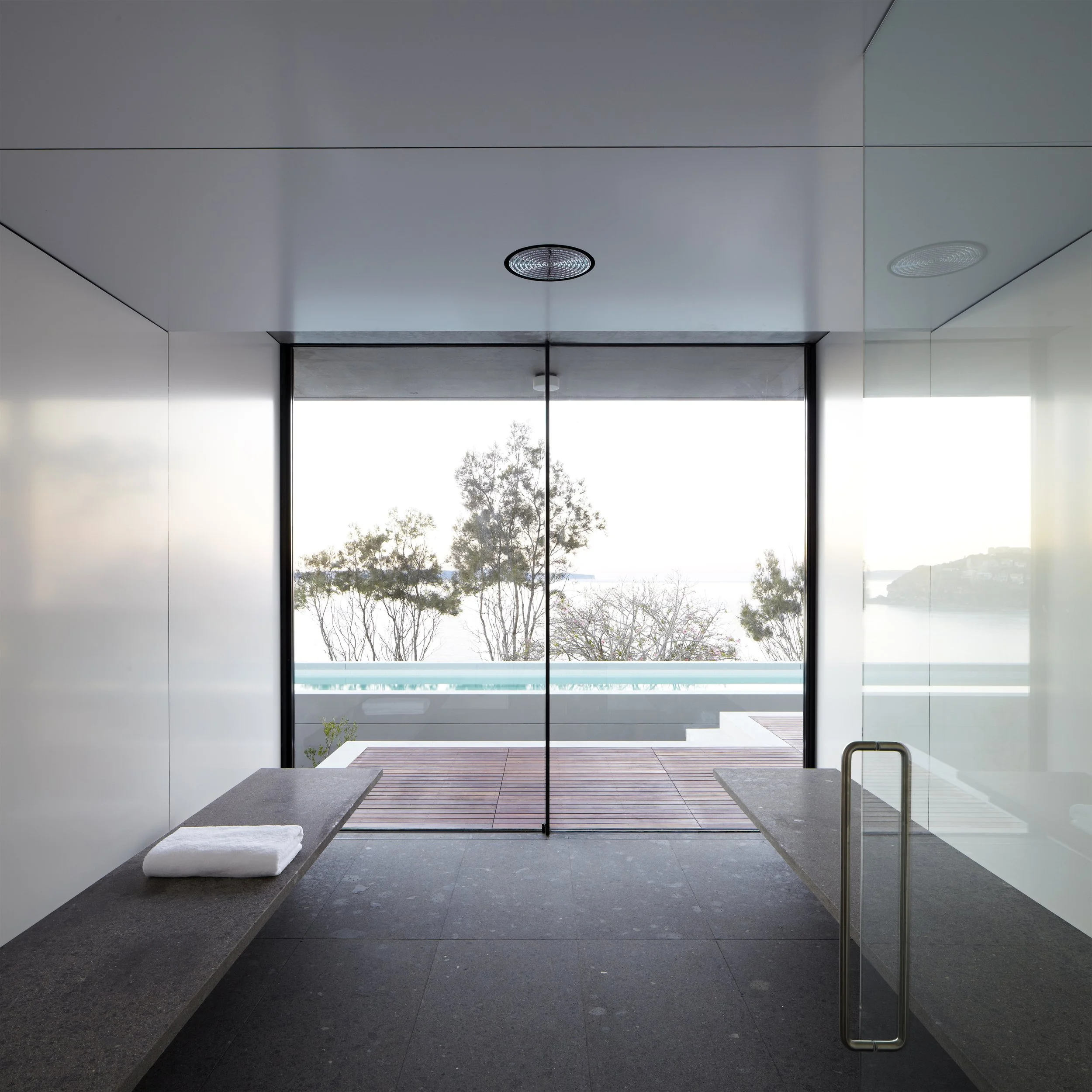 Modern bathroom with a large glass window overlooking trees and water, featuring dark gray stone floors and benches with white towels, rain showerhead, and glass door with metal handle.