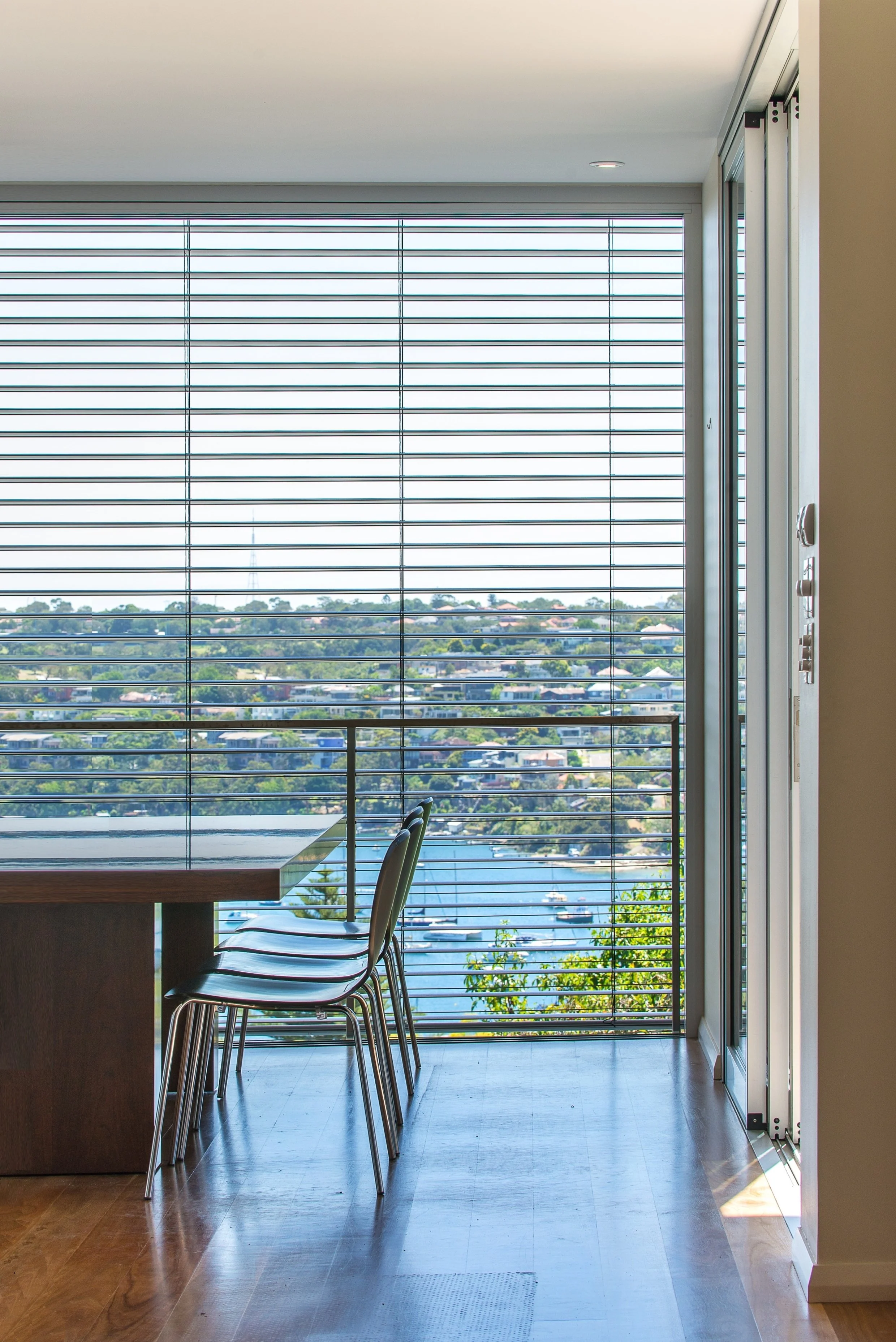 Interior view of a room with a large window with horizontal blinds, overlooking a cityscape with a body of water and boats.