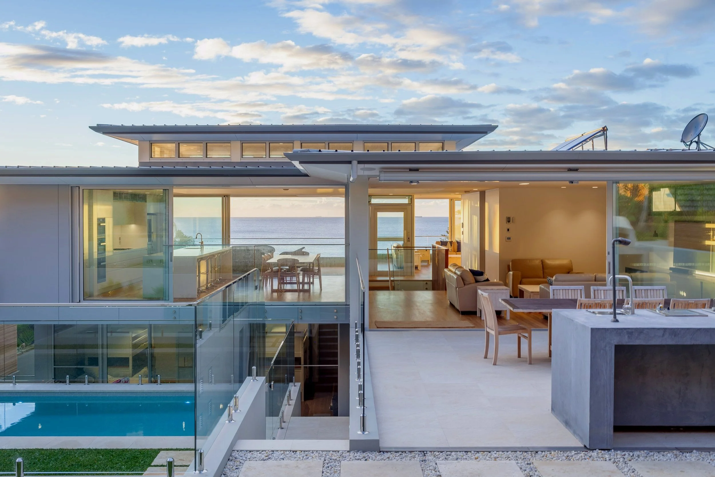 Modern house with outdoor patio, glass railing, and ocean view, interior with living room, dining area, and kitchen visible through large glass sliding doors, pool below, clear sky with scattered clouds.