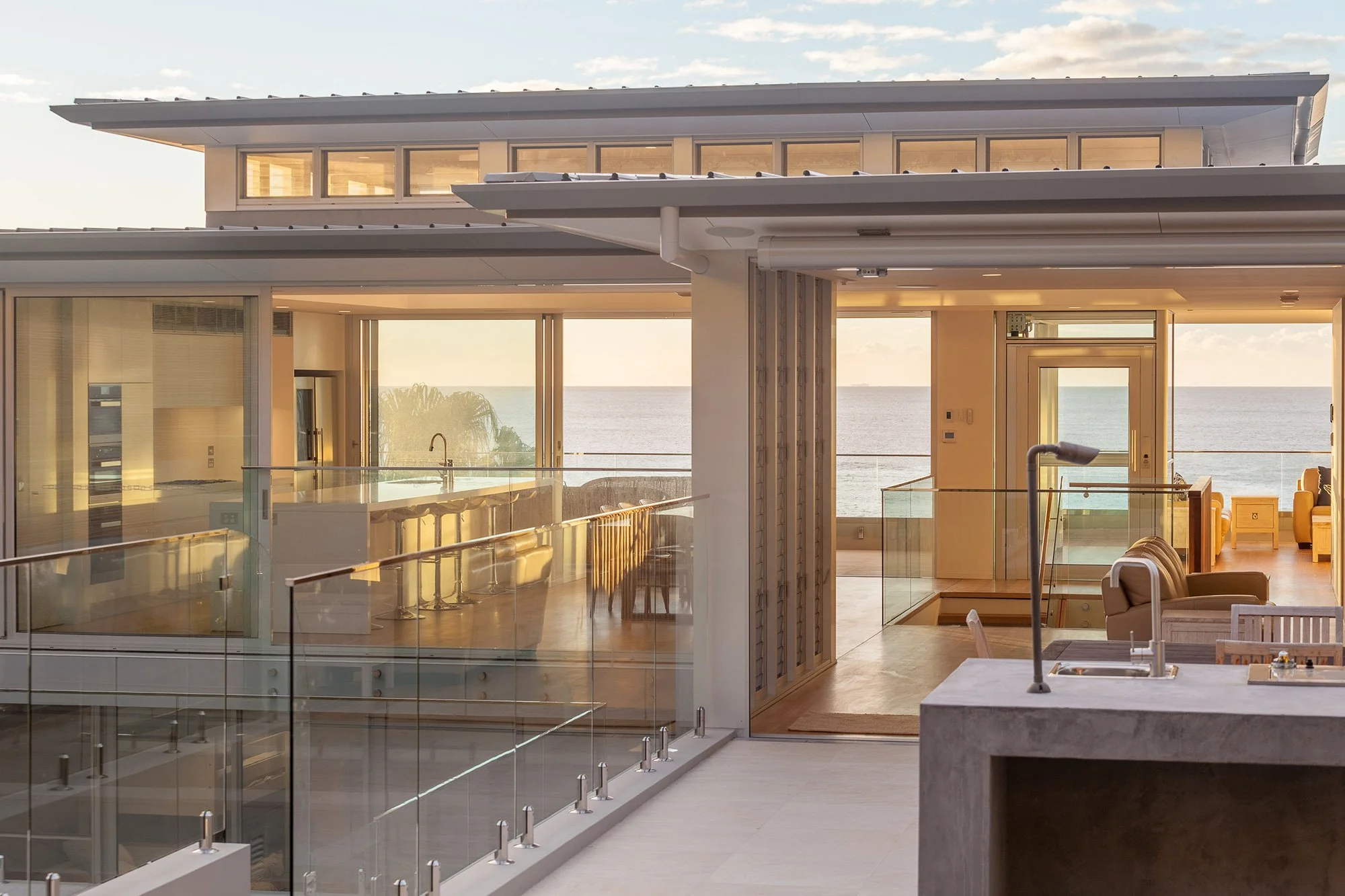 Modern house interior with large glass windows overlooking the ocean, with visible furniture including couches and lounge chairs, and a kitchen island with a sink.