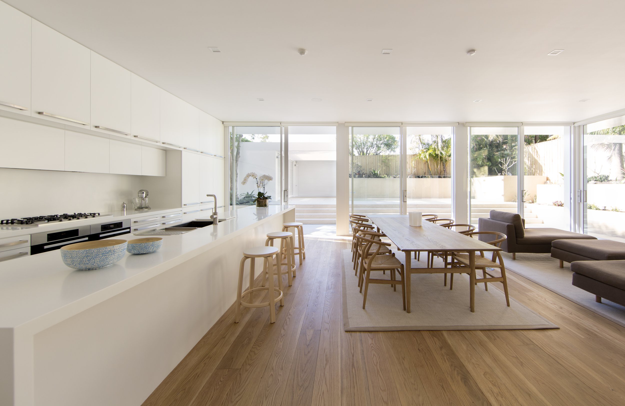A modern, bright open-plan kitchen and dining area with large windows, white cabinetry, a wooden dining table, and outdoor view.