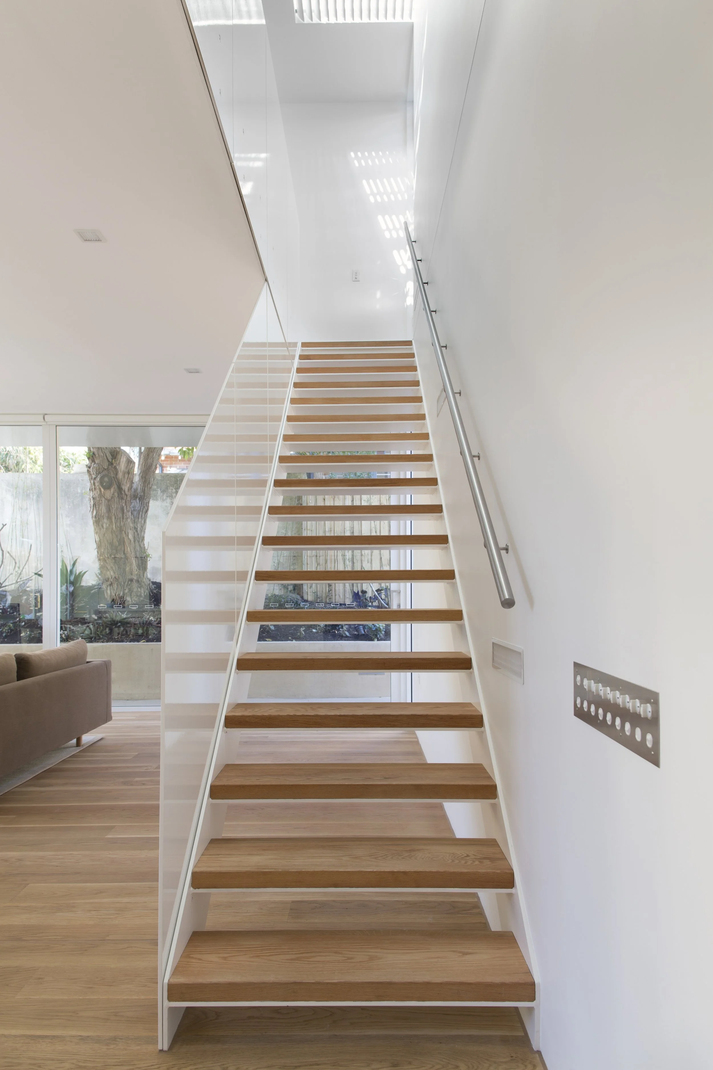 Interior view of a modern staircase with wooden steps and white side supports, stainless steel handrail, and a large window showing trees outside.
