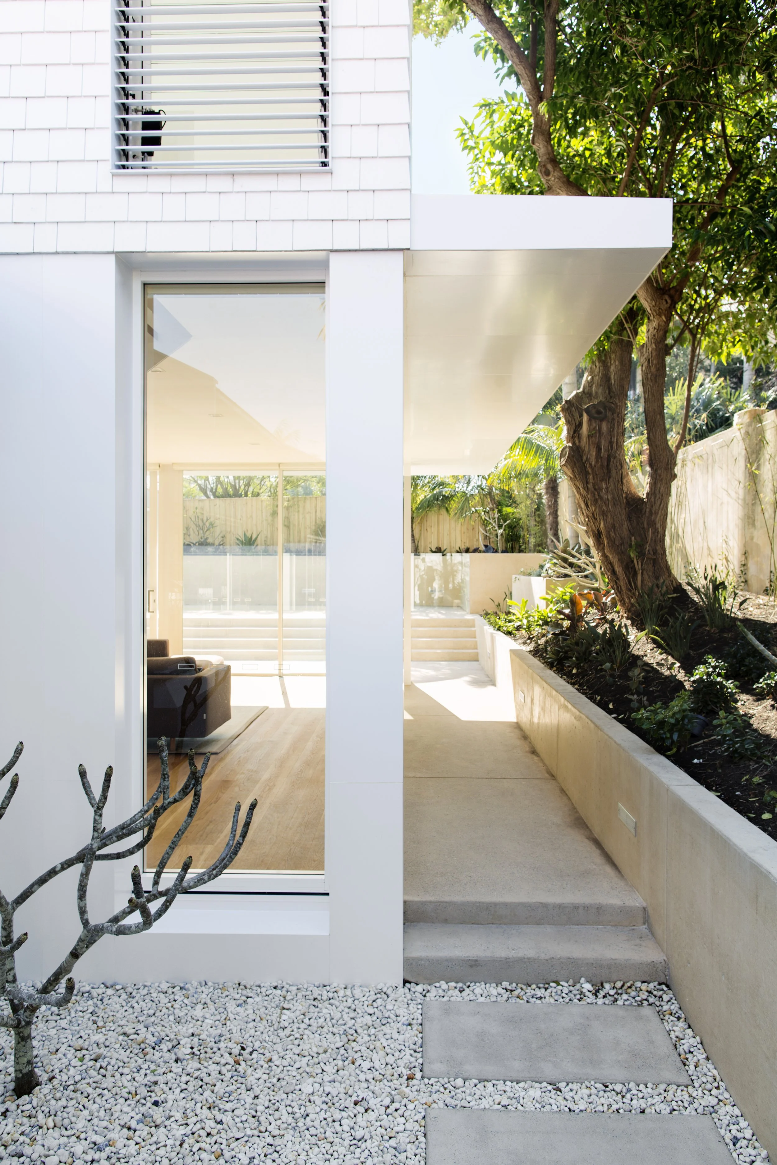 Modern house exterior with large glass door, lush greenery, and a small tree in a landscaped yard with white gravel, stepping stones, and concrete pathway.
