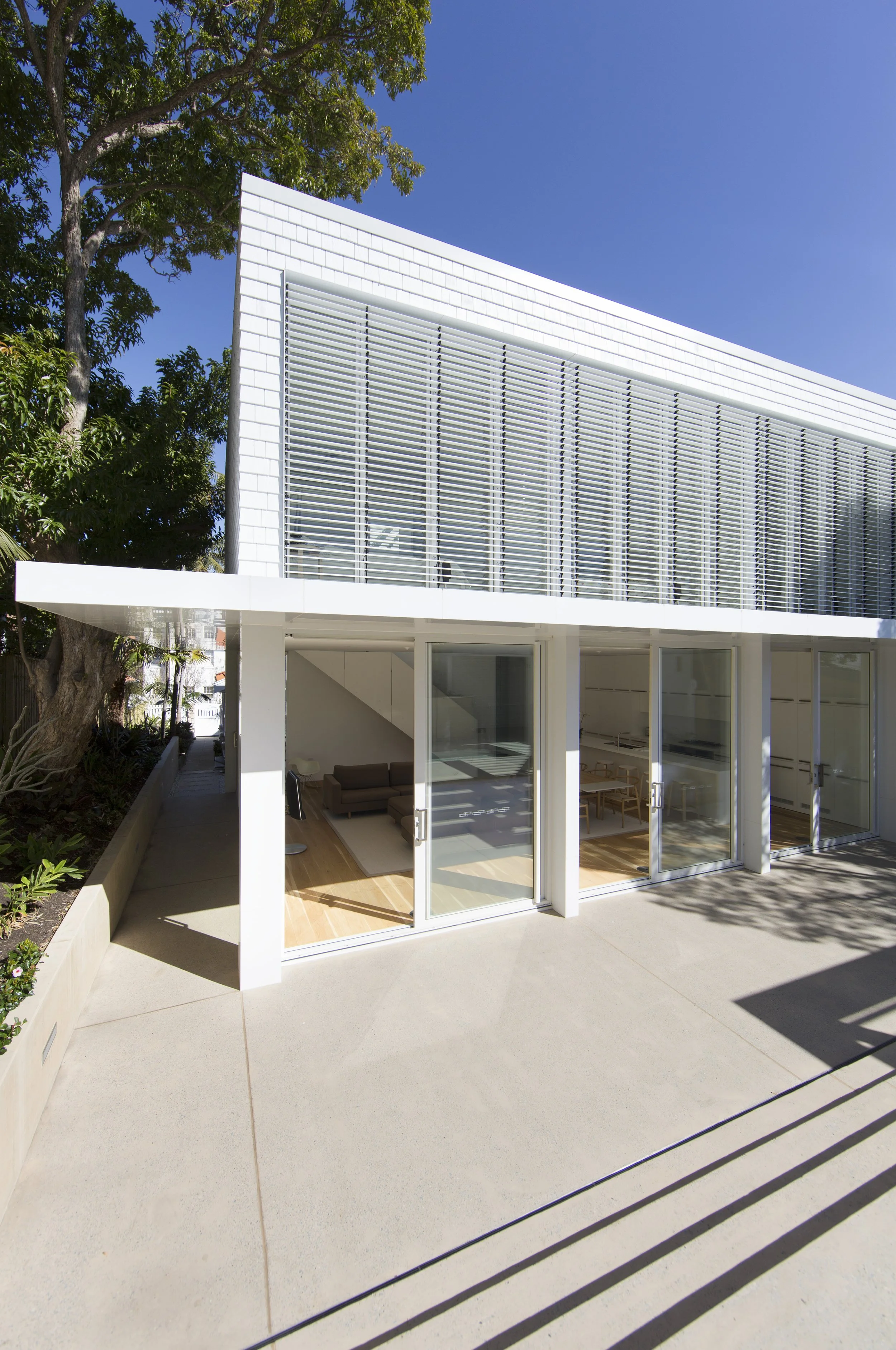 Modern white building with large sliding glass doors and windows, surrounded by trees and a concrete patio, under a clear blue sky.