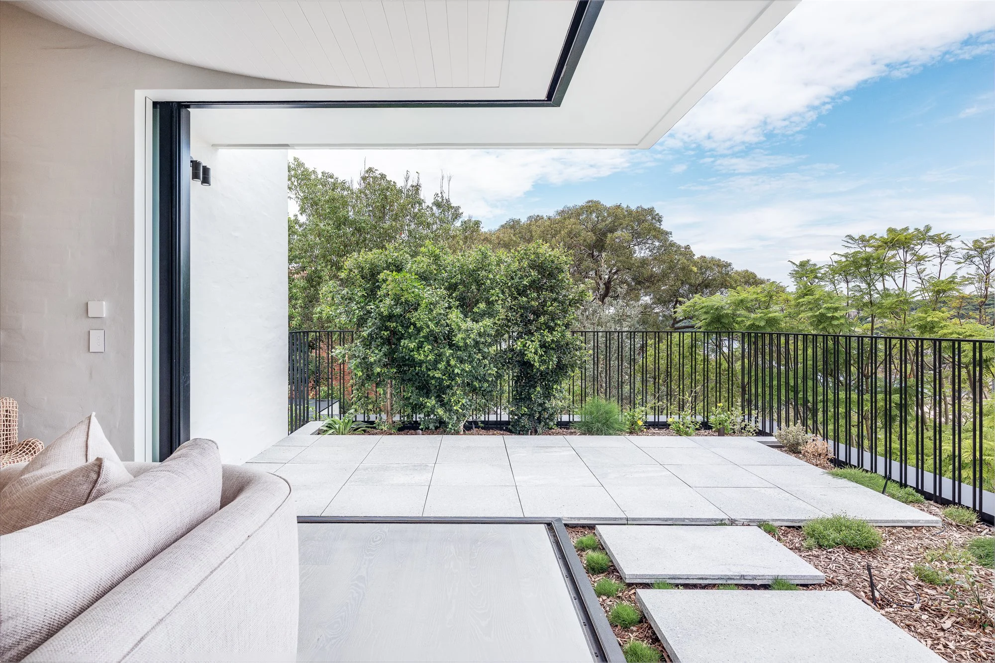 Modern open balcony with a sofa, gray flooring stones, black metal railing, and view of green trees and blue sky.