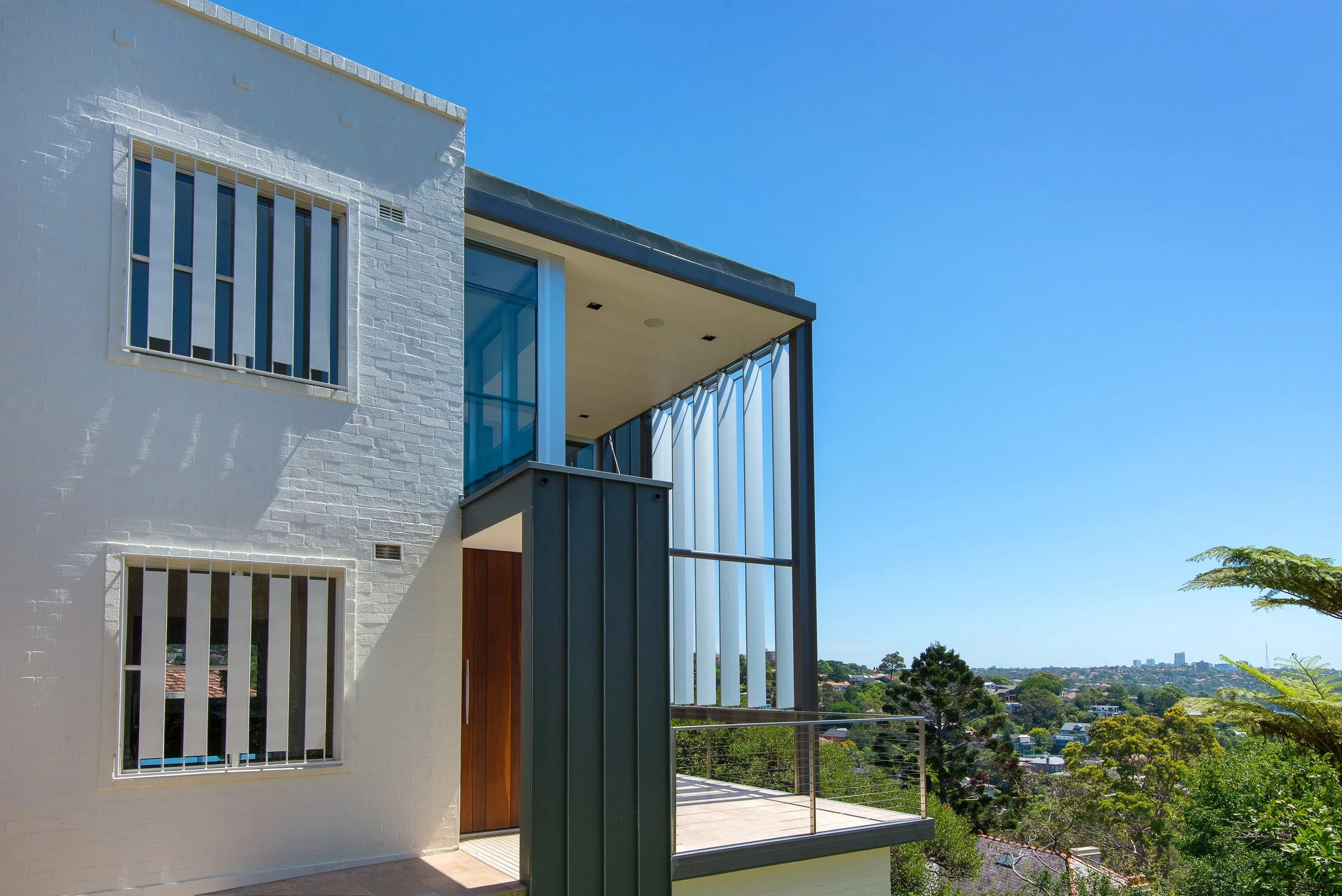 Modern house exterior with white brick walls, large windows with security bars, a wooden door, and a balcony with glass and metal railings, overlooking a green landscape and blue sky.