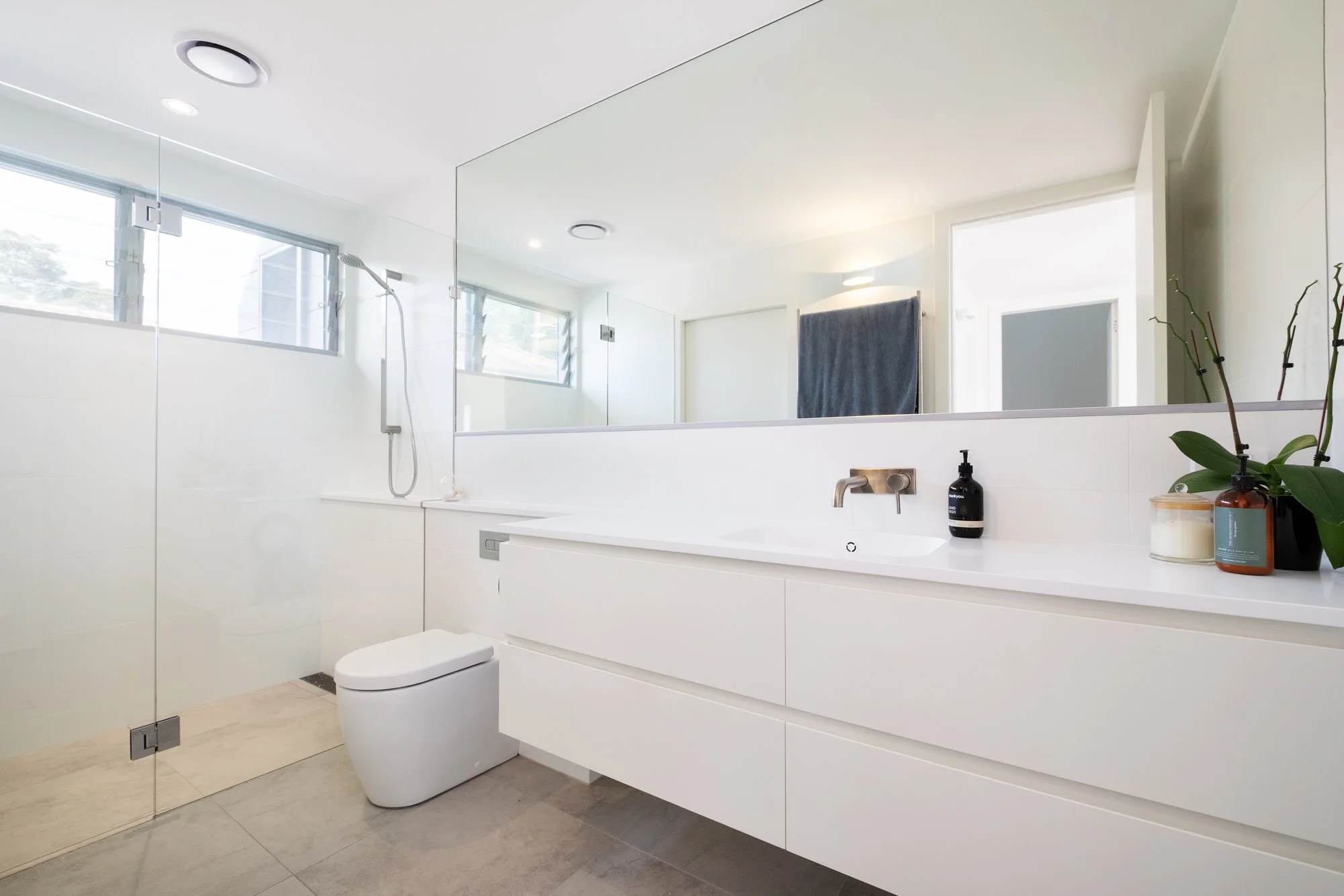 Modern bathroom with white vanity and large mirror, shower area with glass partition, window, and decorative plants.