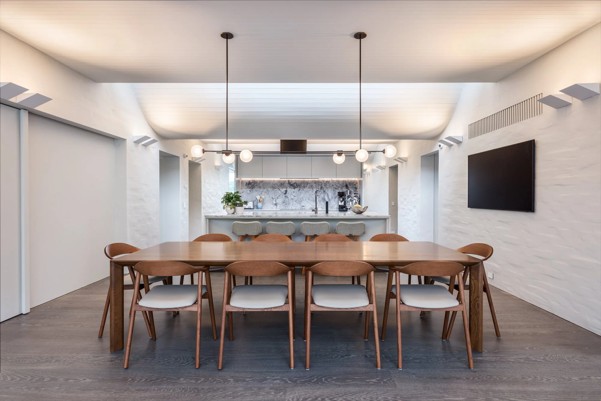 Modern dining room with a wooden table surrounded by eight matching wooden chairs with white cushions, and a kitchen with a bar counter and six stools in the background.