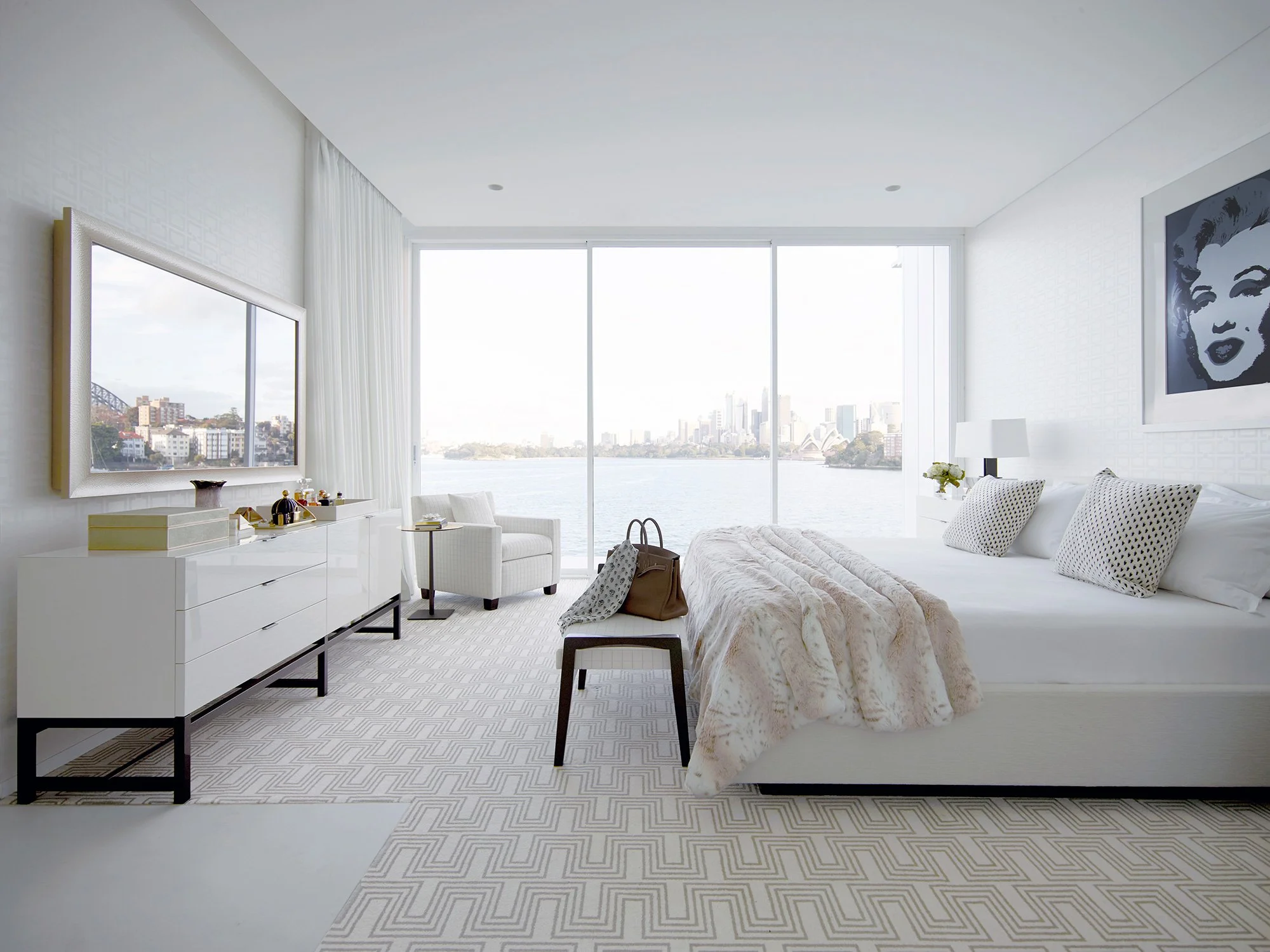 Interior of a modern, white bedroom with a large window overlooking a body of water and city skyline, featuring a bed with pillows and a blanket, a white dresser with a mirror, a chair, and artwork on the wall.