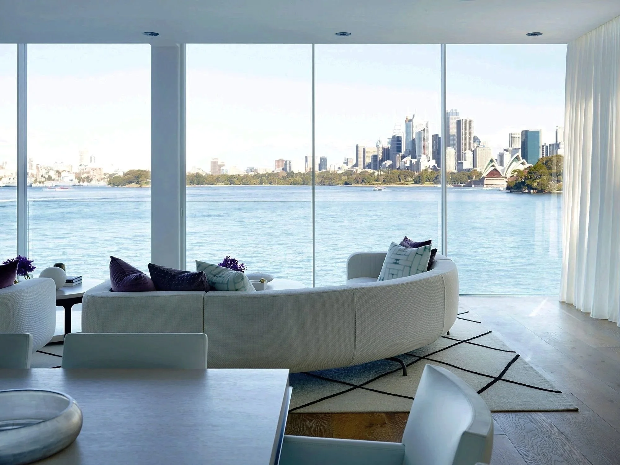 Modern living room with large floor-to-ceiling windows overlooking a body of water and a city skyline with skyscrapers, a framed sofa with pillows, a coffee table with flowers, and a patterned rug.