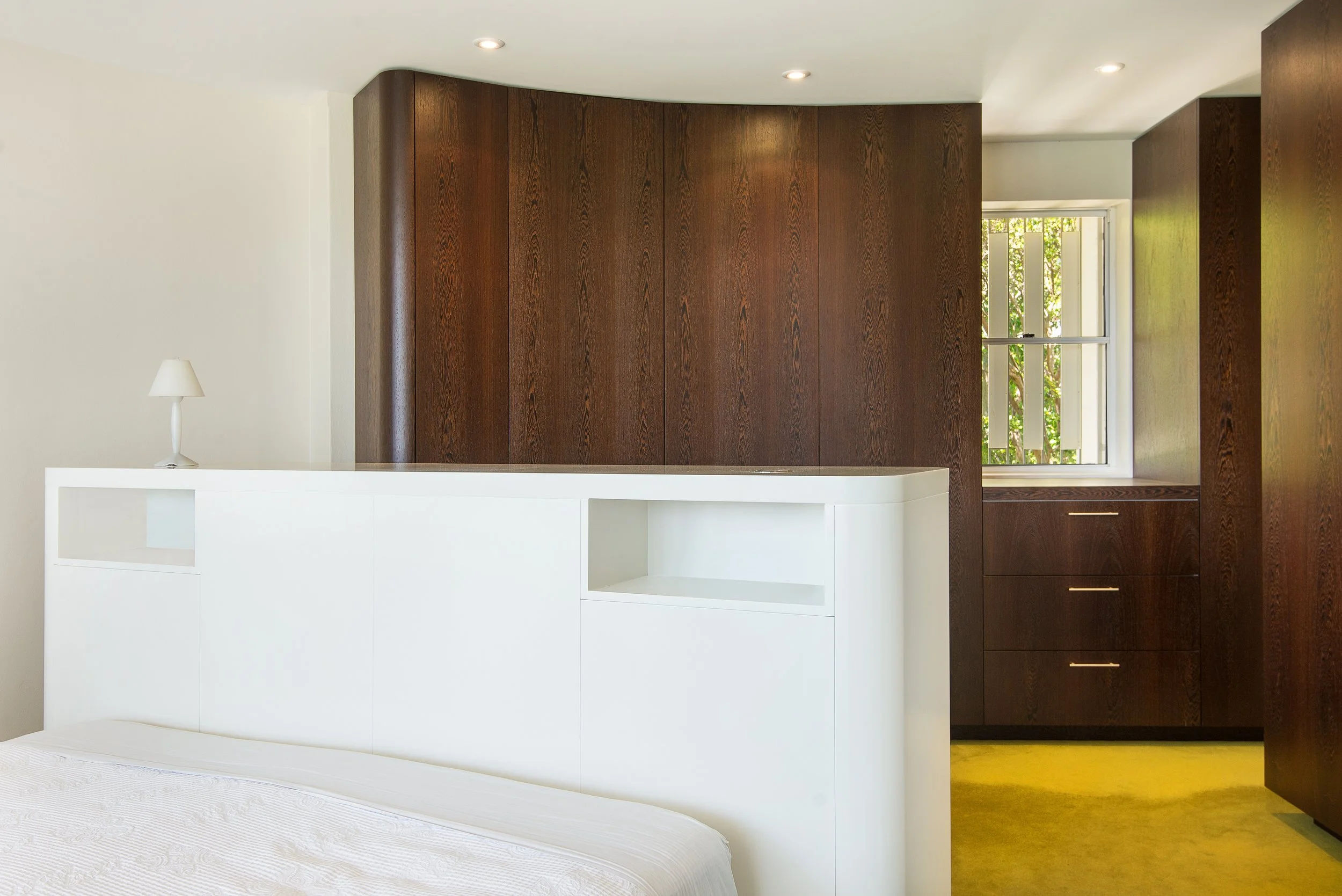 Interior of a bedroom with a white bed frame, a small white lamp on the bed's headboard, dark wood built-in cabinets, a window with white blinds, and a yellow carpet.