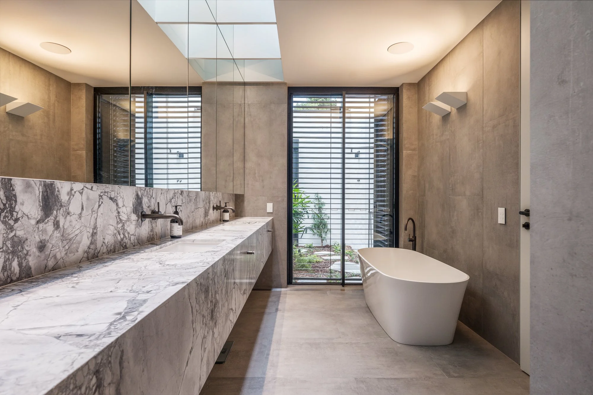 Modern bathroom with a double marble vanity, two sinks, a freestanding bathtub, large window with blinds, and neutral-toned walls.