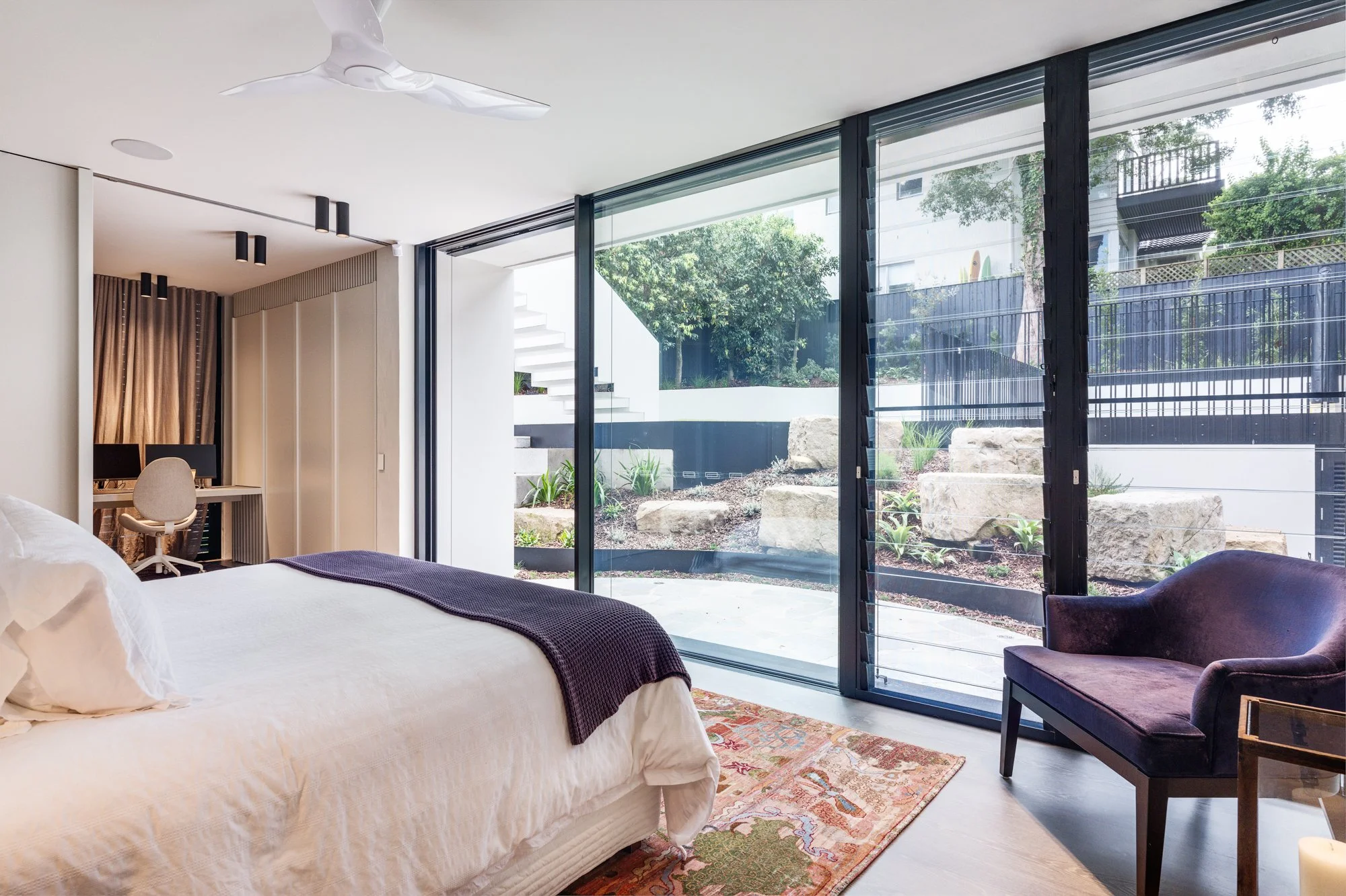 Modern bedroom with large glass sliding doors opening to outdoor garden with rocks and plants, a bed with white bedding and purple blanket, a purple armchair, a small table, a ceiling fan, and a desk with computer.