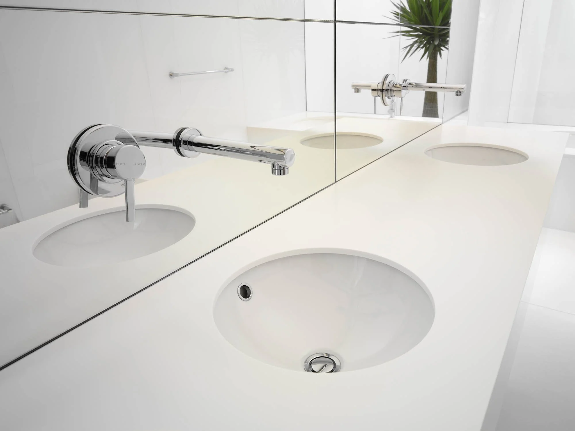 A modern white bathroom countertop with two round sinks, a large mirror, and a faucet, with a potted plant visible in the reflection.