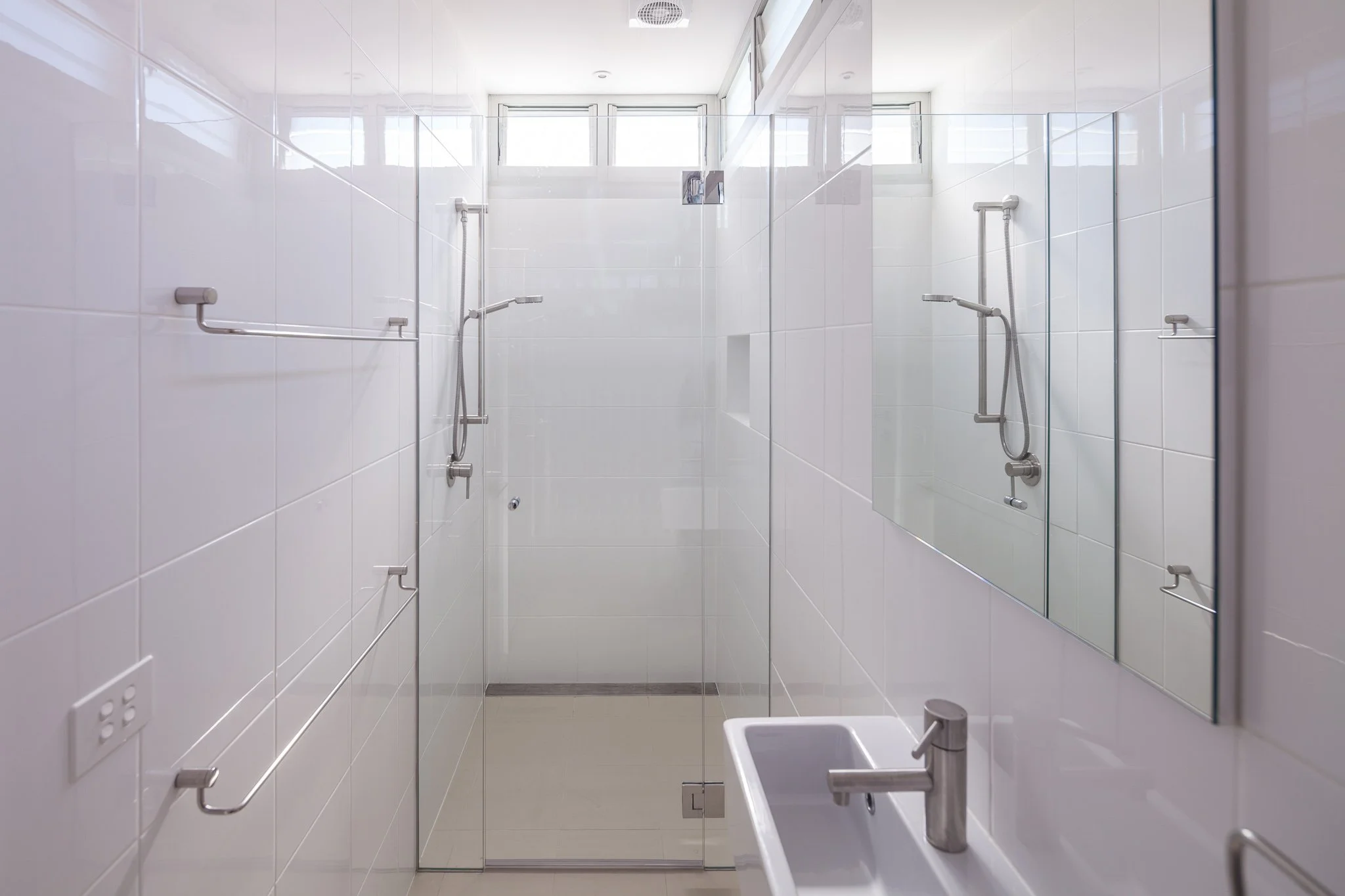 Modern white shower with a glass door, wall-mounted showerheads, and a white sink with a silver faucet in a bright bathroom.