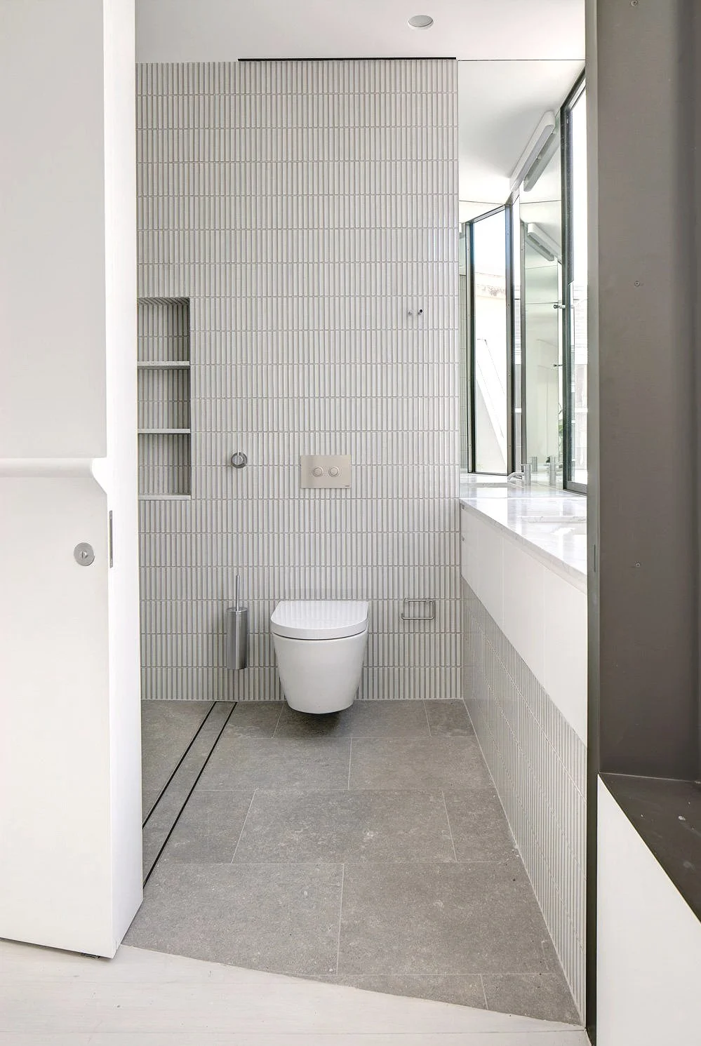 Modern bathroom with a wall-mounted toilet, gray floor tiles, and large windows letting in natural light.