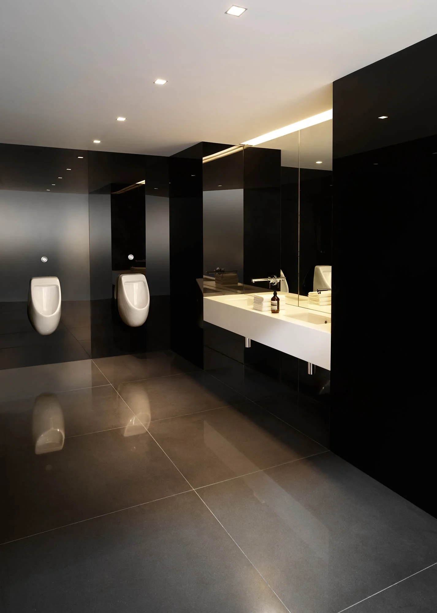 Modern men's restroom with black walls, white urinals, a white sink, and large reflective floor tiles.