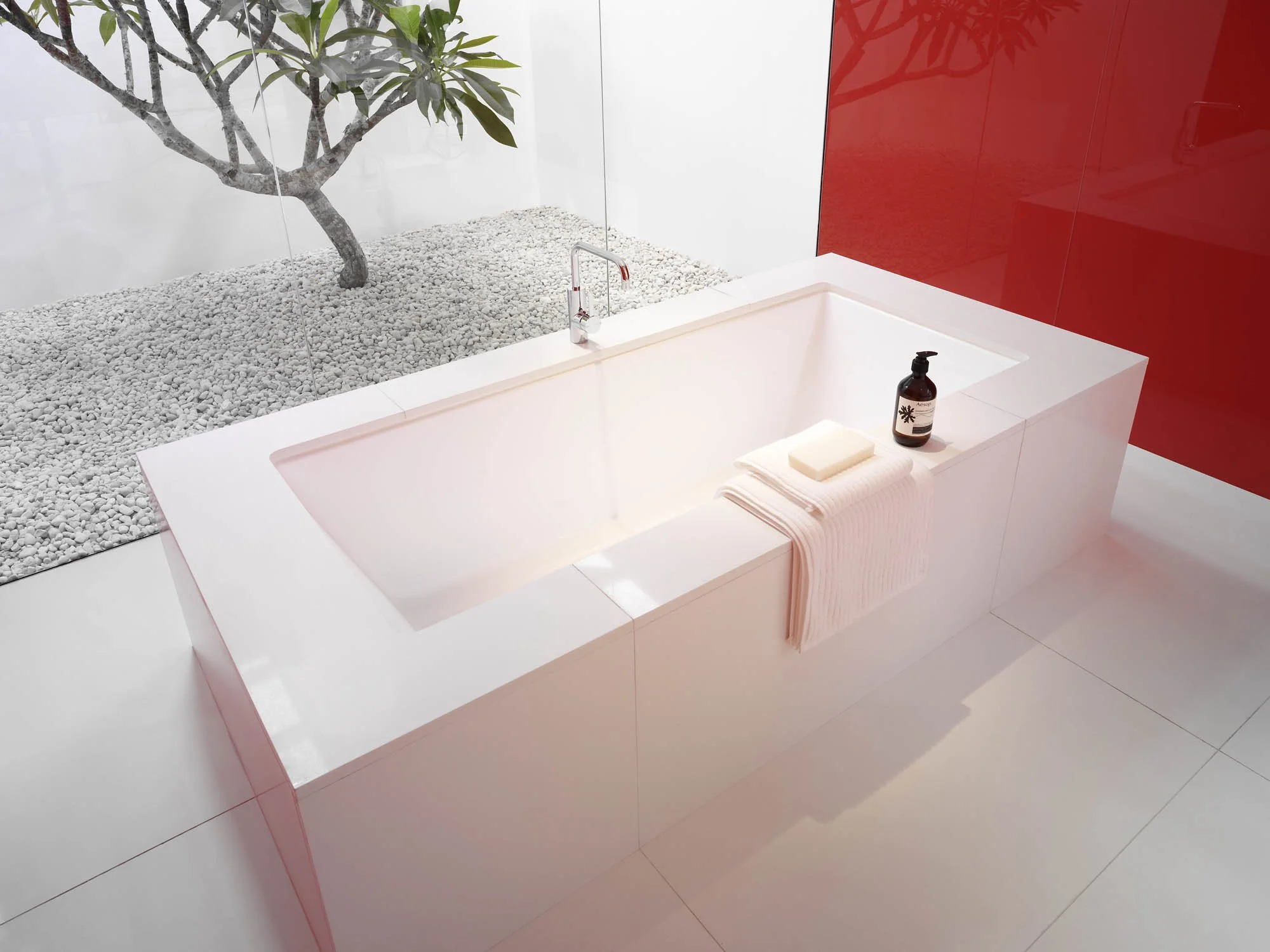 Modern bathroom with a rectangular white bathtub, a towel hanging on the side, and a bottle of soap or lotion on the edge. There's a soap dish on the towel. Behind the tub is a large window with a view of a small tree and white gravel outside, and a 
