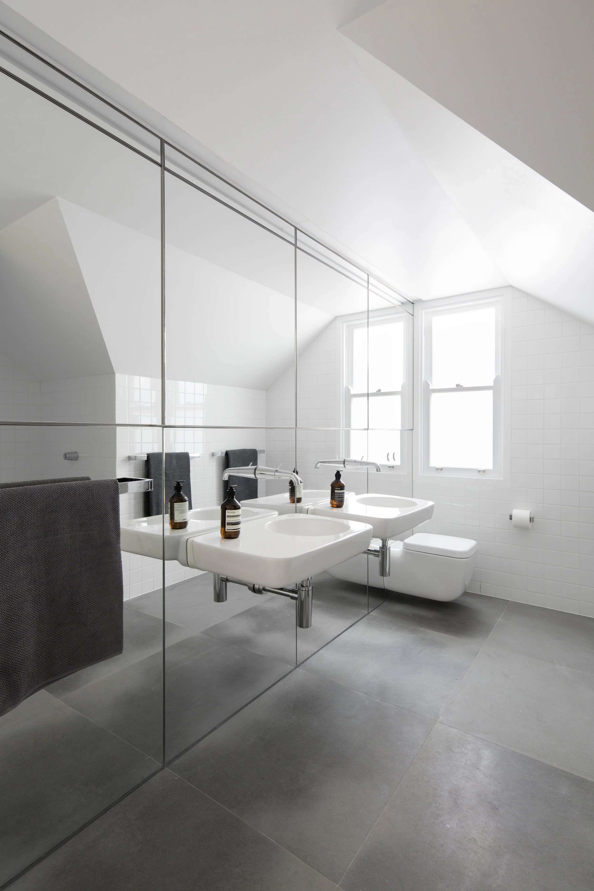 A modern bathroom with a large mirror, two sinks, and a toilet, featuring white tiles, gray floor tiles, and two windows letting in natural light.