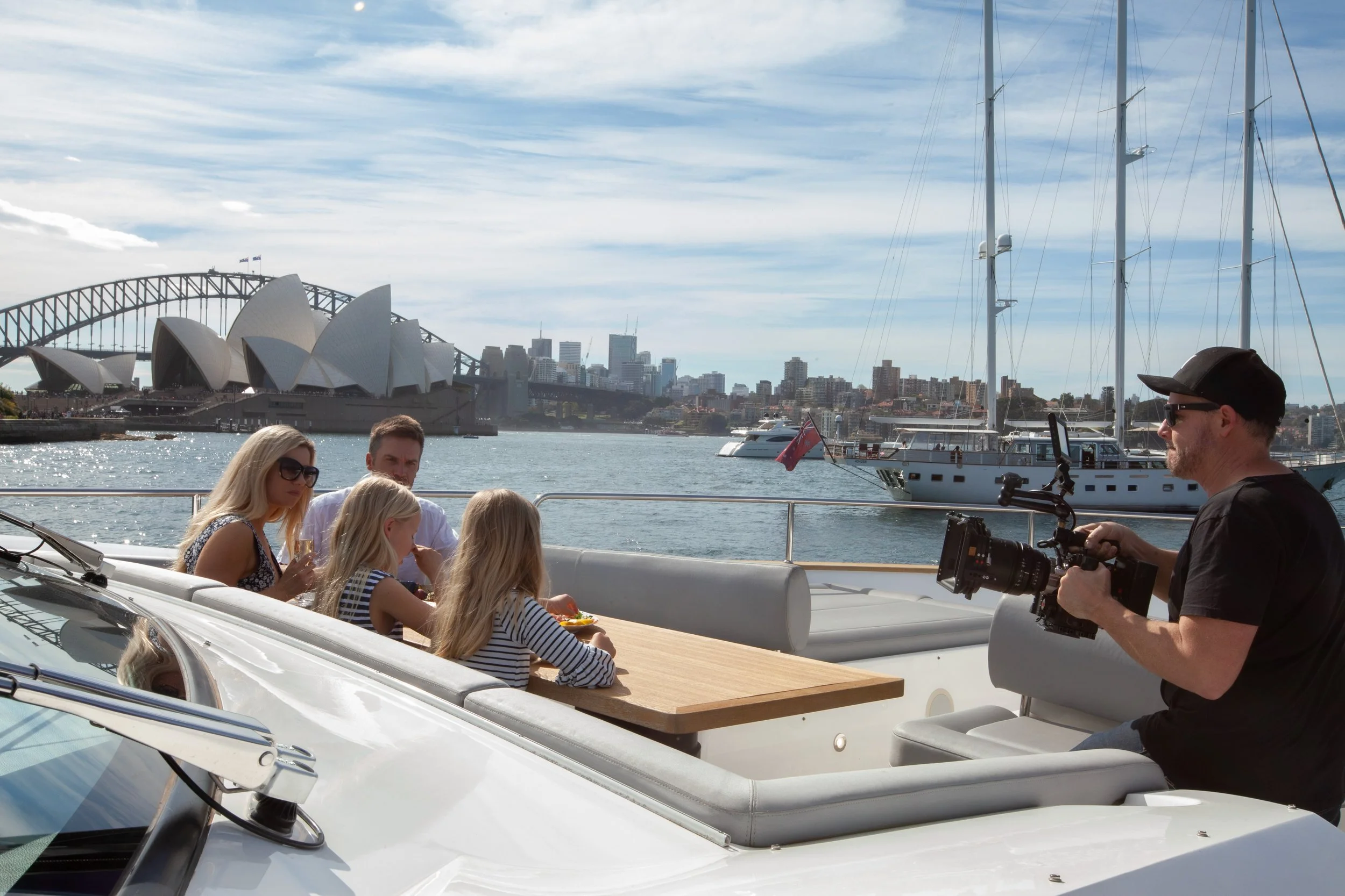 A family of four sitting on a yacht with a filmer on the right capturing video, with the Sydney Opera House and city skyline in the background during the day.