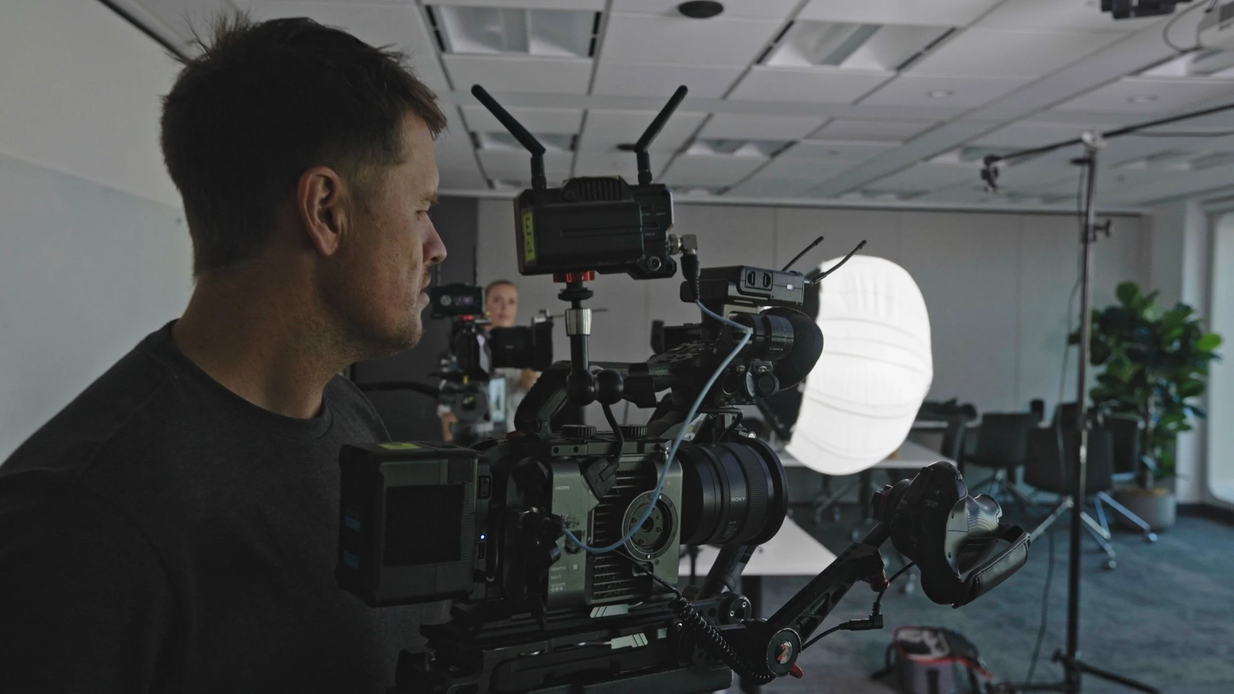 A man operating a professional video camera in an indoor studio setting, with a large white light diffuser and a woman in the background at another camera setup.