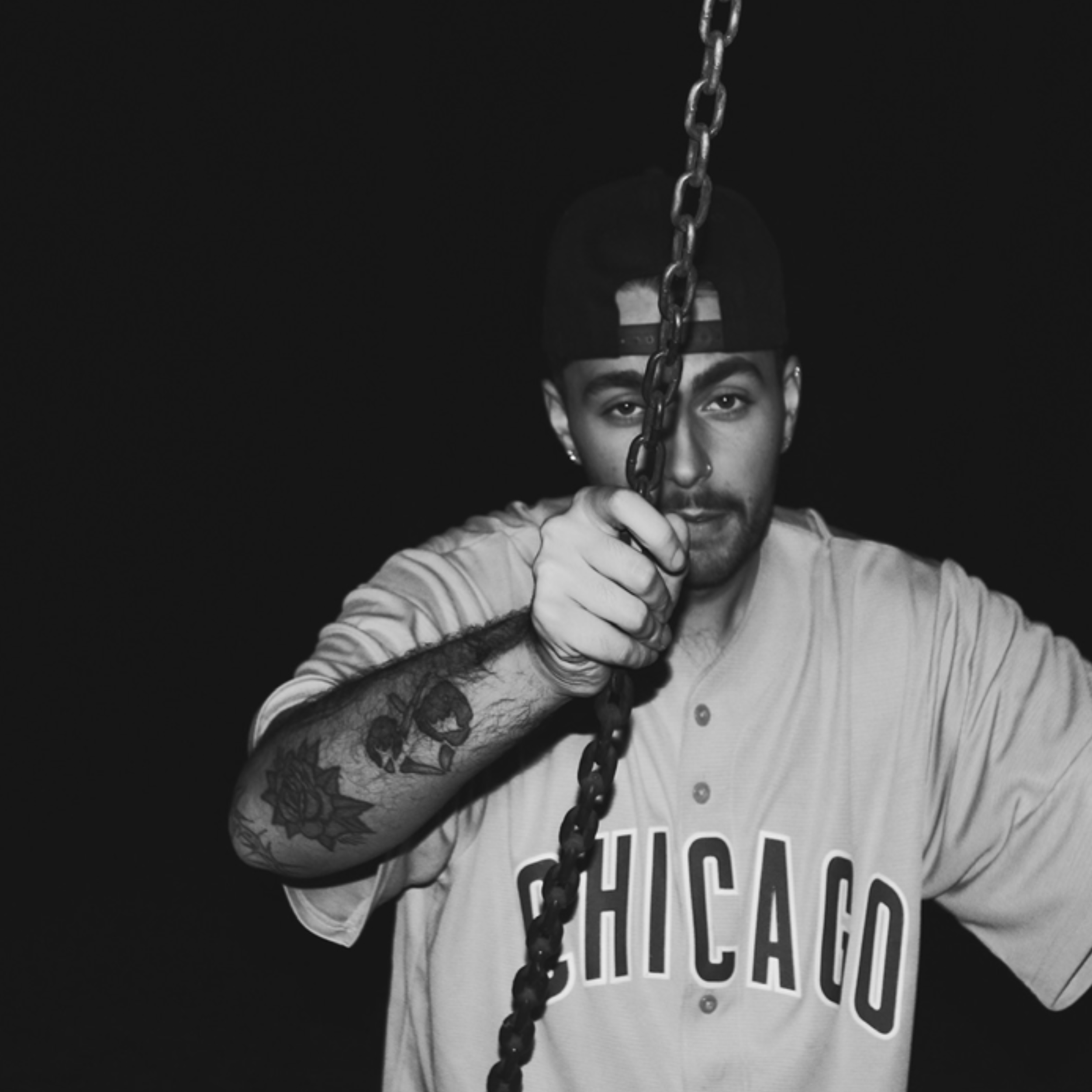 A young man wearing a baseball jersey with 'Chicago' written on it, a cap worn backward, and tattoos on his arm, holding a chain in front of his face against a dark background.