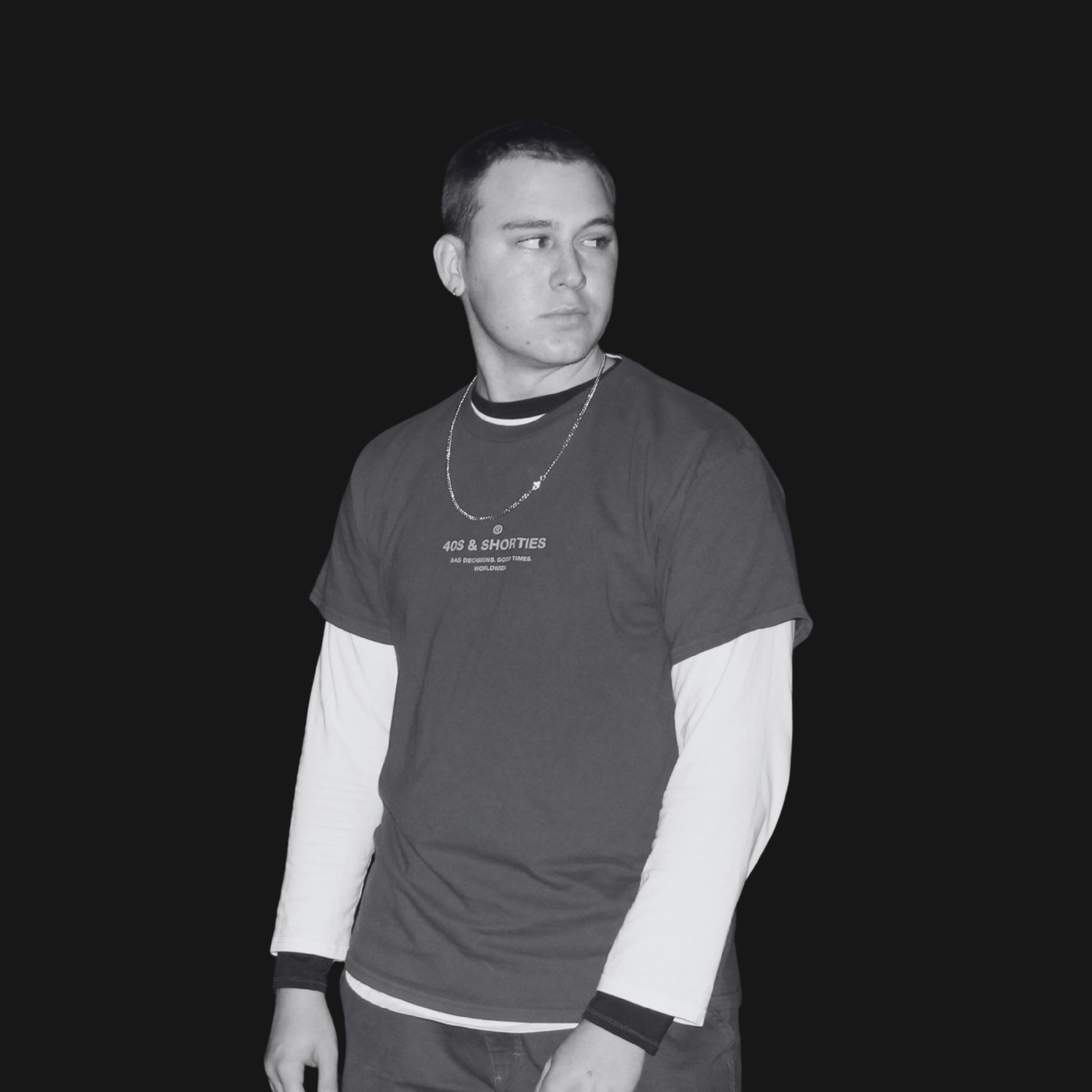 Young man with short hair wearing a graphic t-shirt layered over a long-sleeve shirt, standing against a black background, looking to the side.