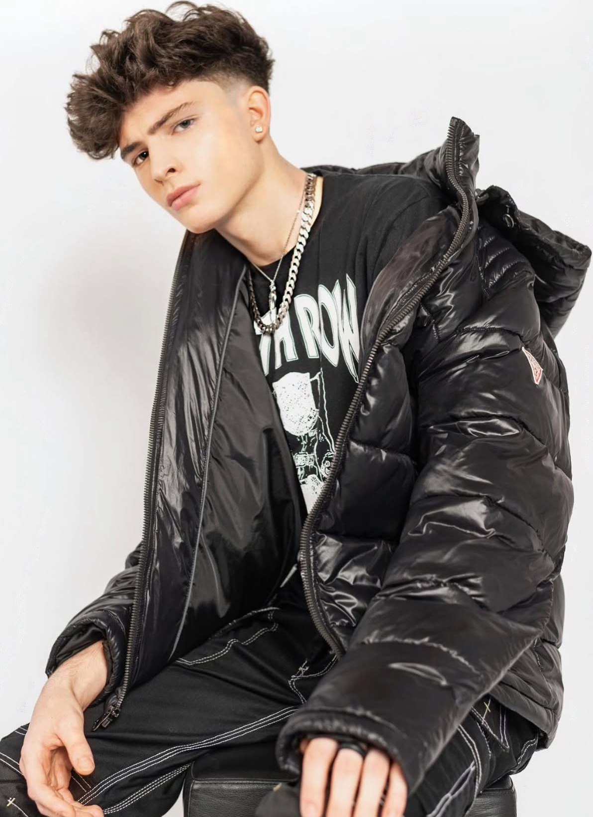 A young man with styled, curly brown hair, wearing earrings, a chain necklace, a black graphic t-shirt, and a shiny black puffer jacket, sitting against a plain white background.