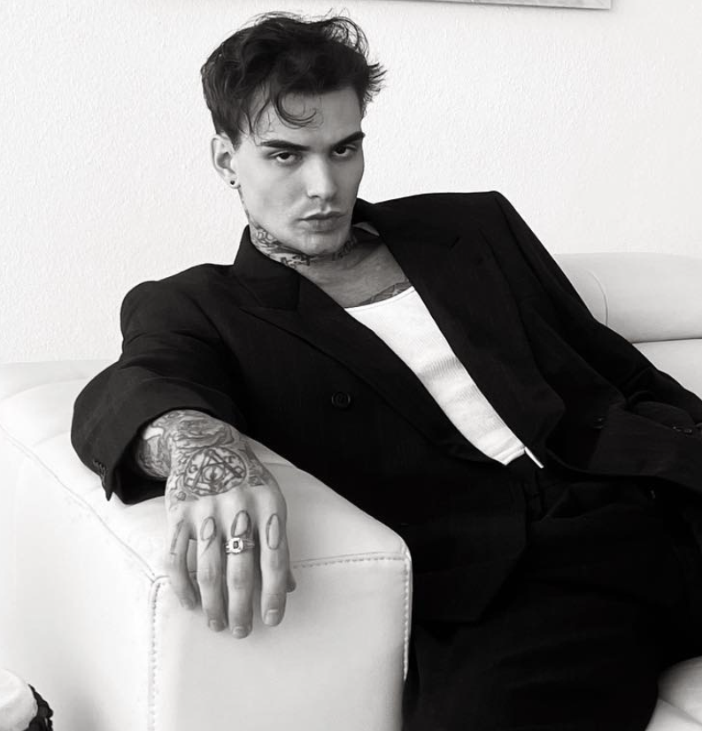 A young man with short dark hair, tattoos on his neck and left hand, wearing earrings and rings, dressed in a black blazer over a white shirt, sitting on a white sofa with a serious expression, in a room with a plain wall.