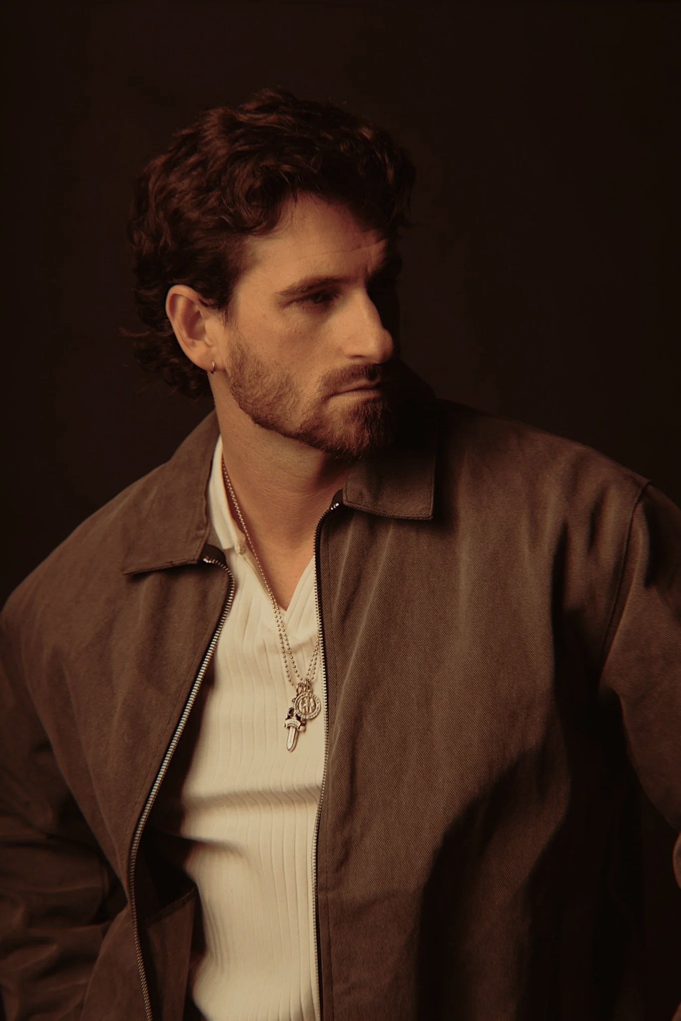 A man with dark, curly hair and a beard, wearing a brown jacket over a white shirt, looking to the left against a dark background.
