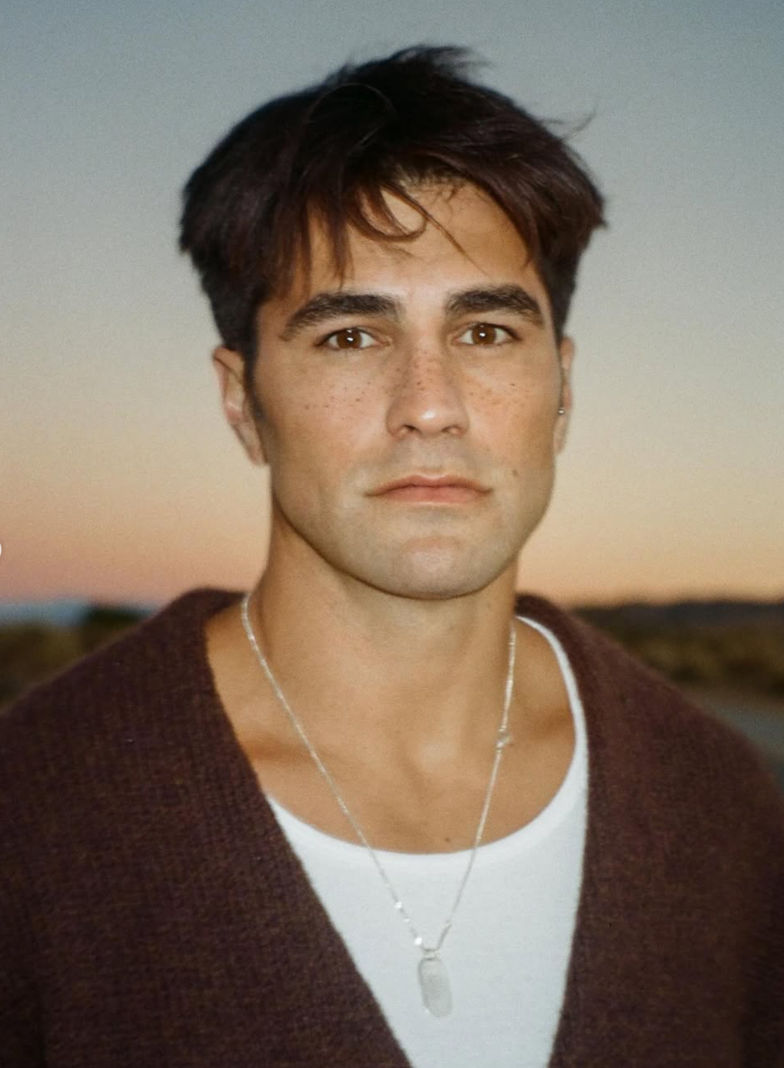 A young man with short dark hair, light skin with freckles, wearing a white shirt, a dark brown cardigan, and a silver necklace, standing outdoors at sunset.