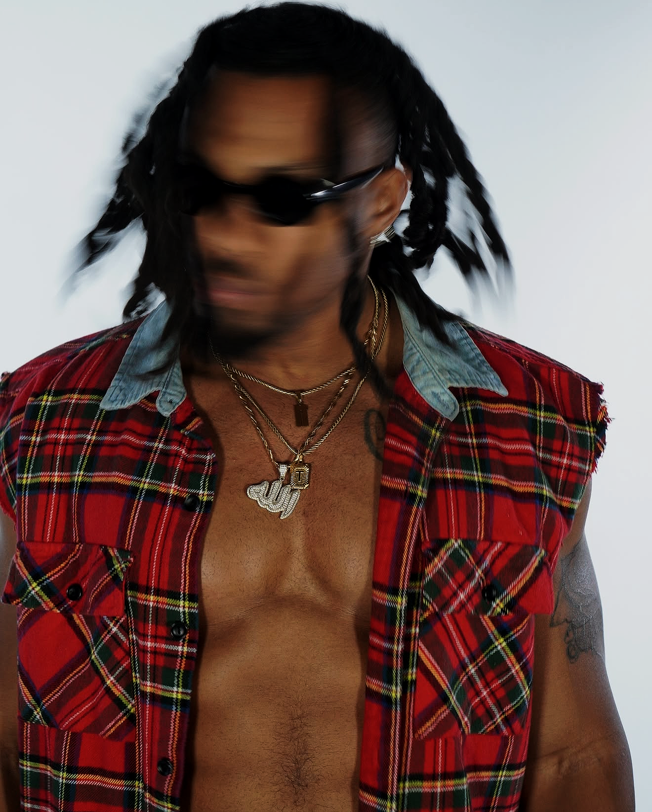 A person with long dreadlocks wearing dark sunglasses, layered gold necklaces, and a sleeveless red plaid vest over a denim shirt, with a white background.