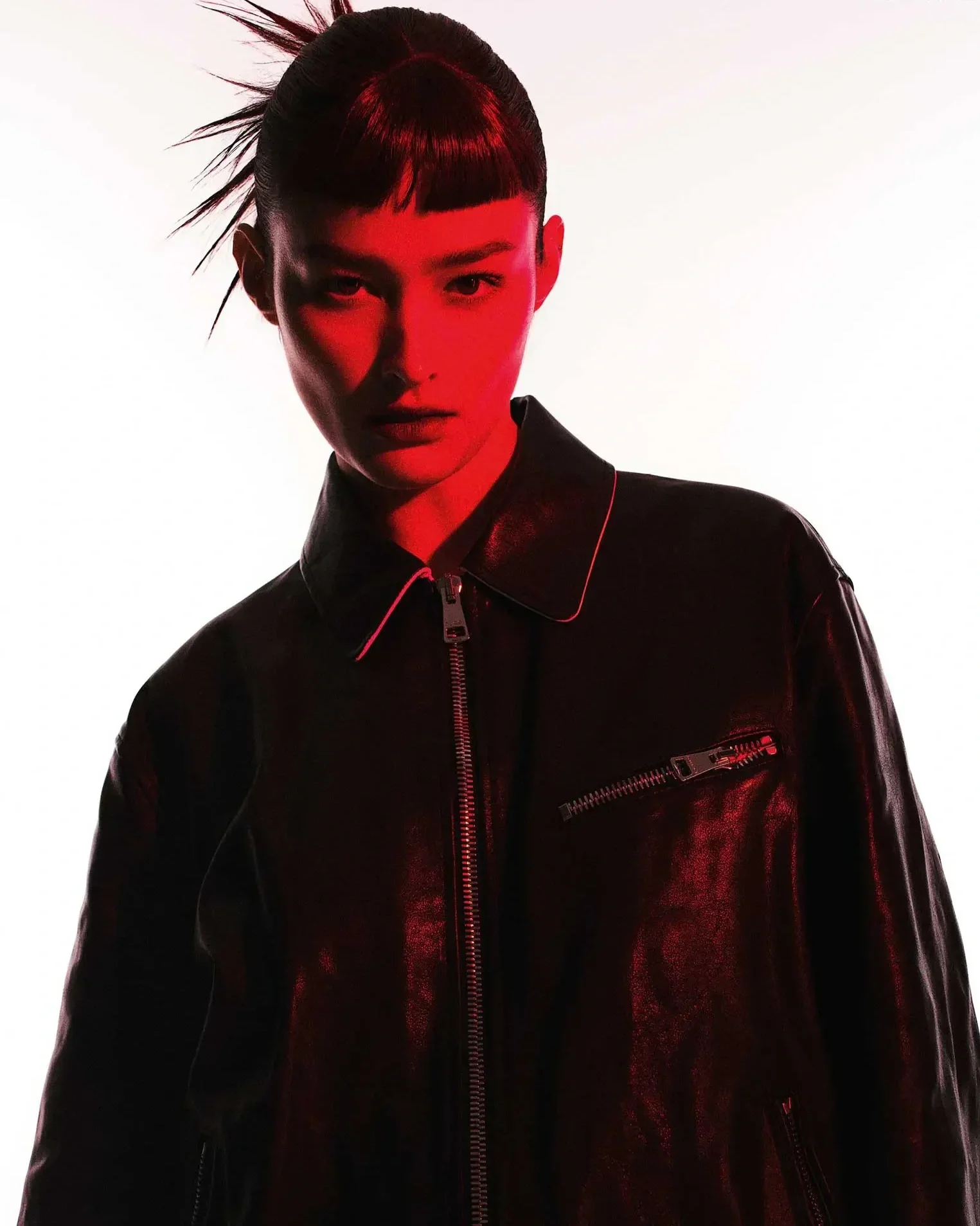 A young woman with a hairstyle featuring spiked and blunt cut bangs, wearing a black leather jacket and illuminated with red lighting, standing against a white background.