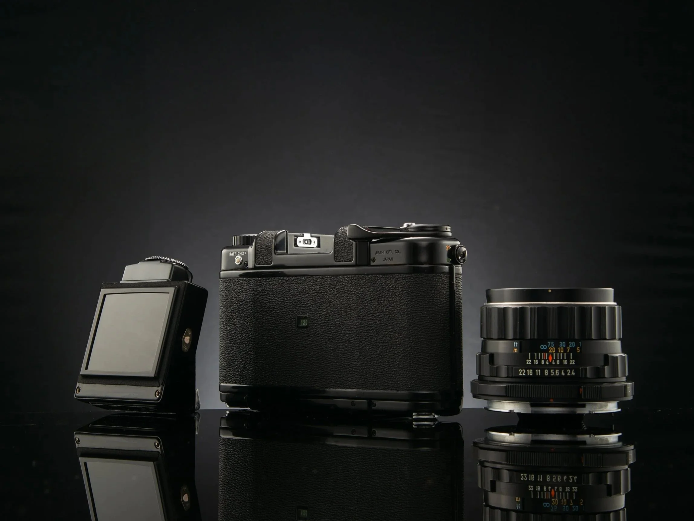 A vintage film camera with a detachable lens and a digital camera display on a reflective black surface.