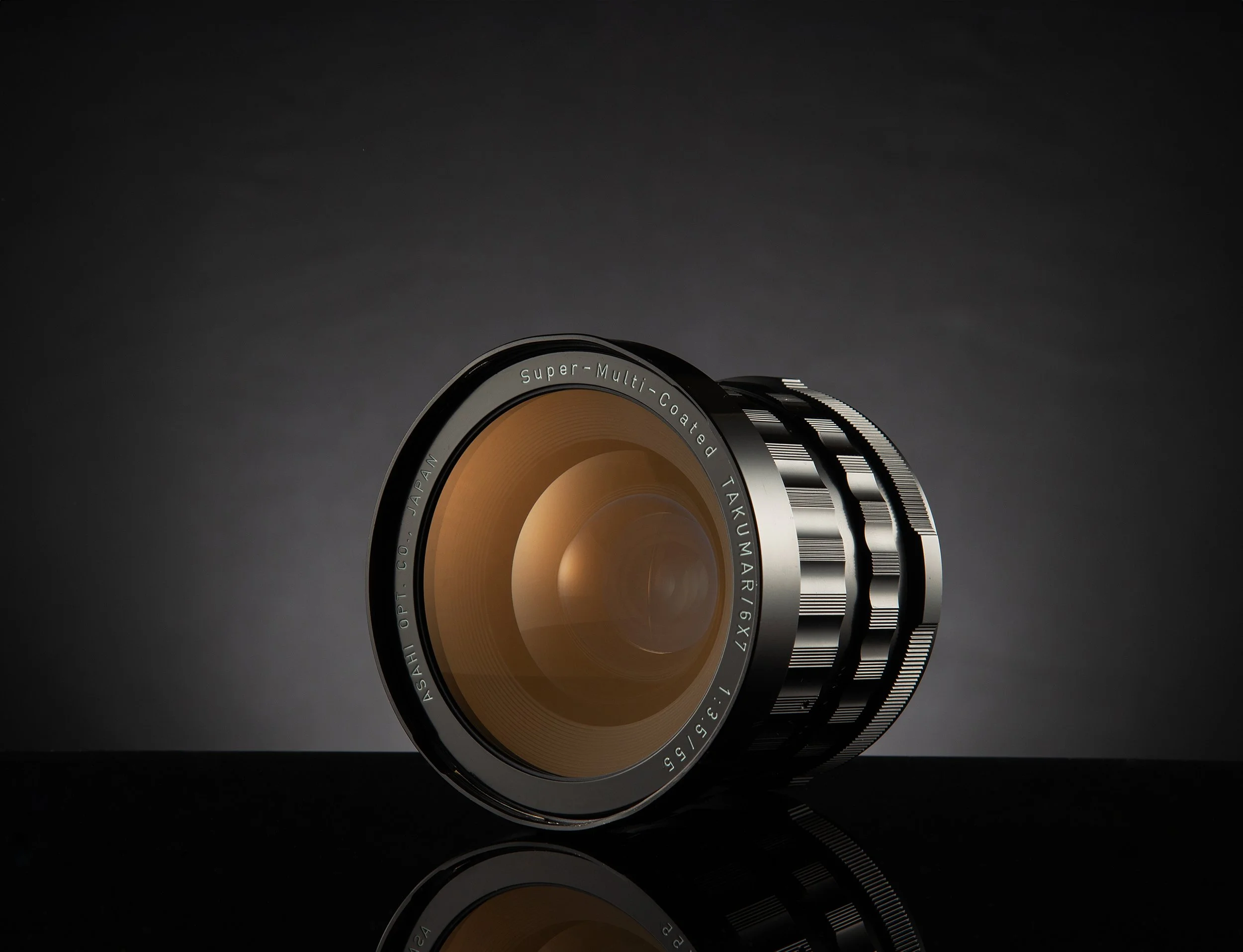 Camera lens with a black and gold design, placed on a reflective surface against a dark background.