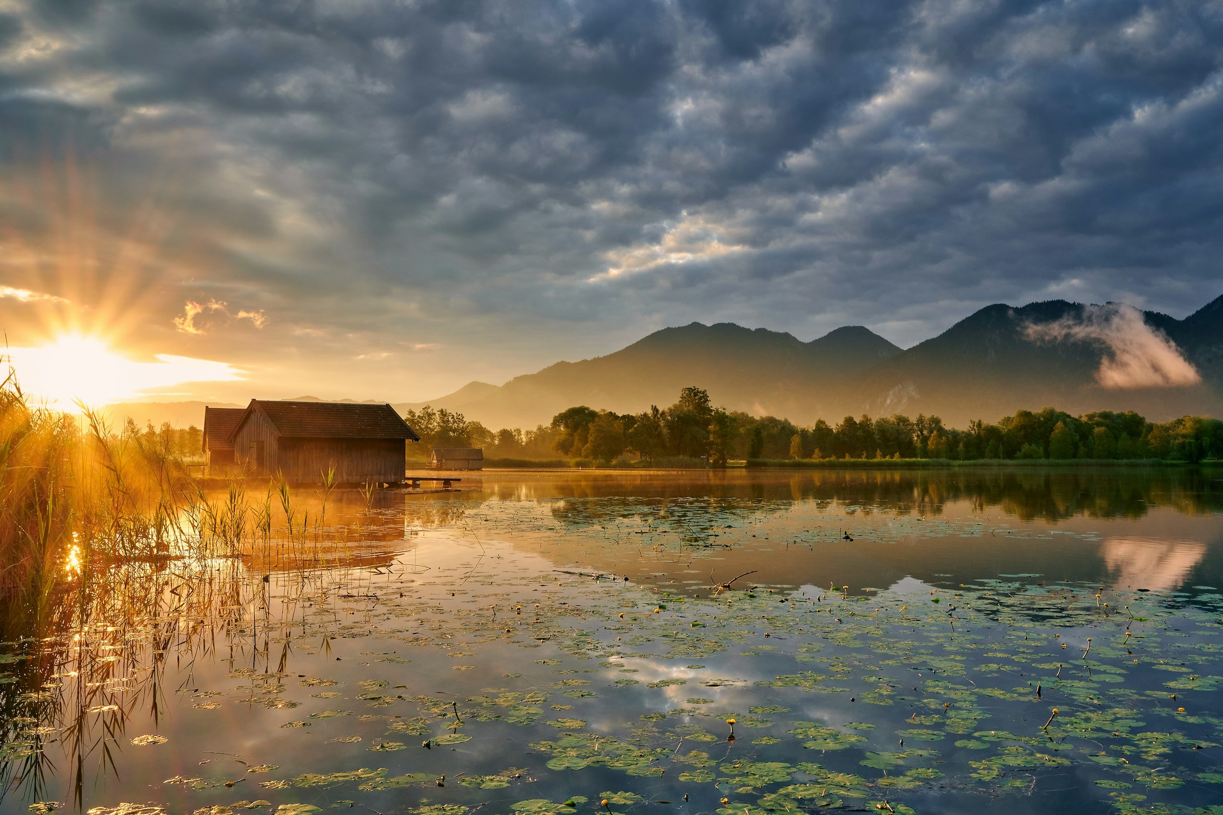 Sunset over a calm lake with lily pads, wooden houses on stilts, and mountains in the background under a cloudy sky.