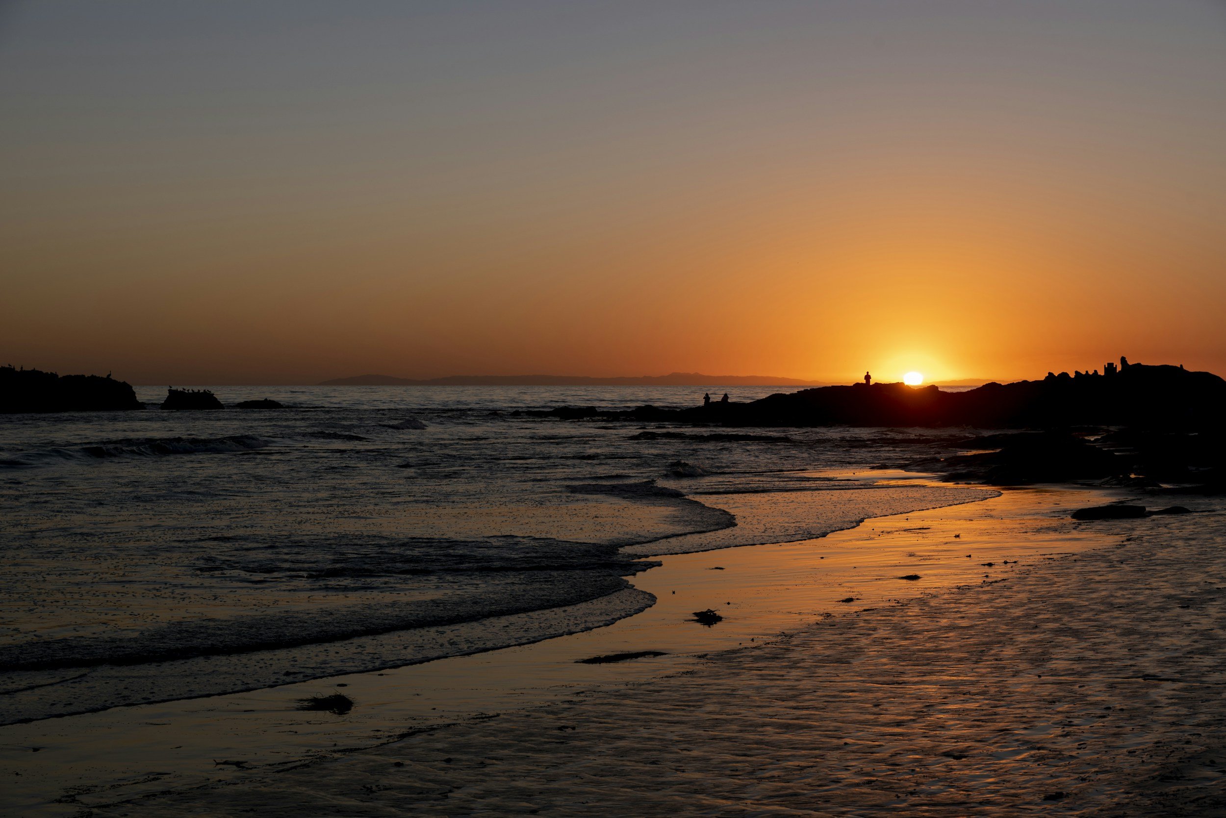 Sunset at the beach with silhouettes of rocks and people, gentle waves, and a colorful sky