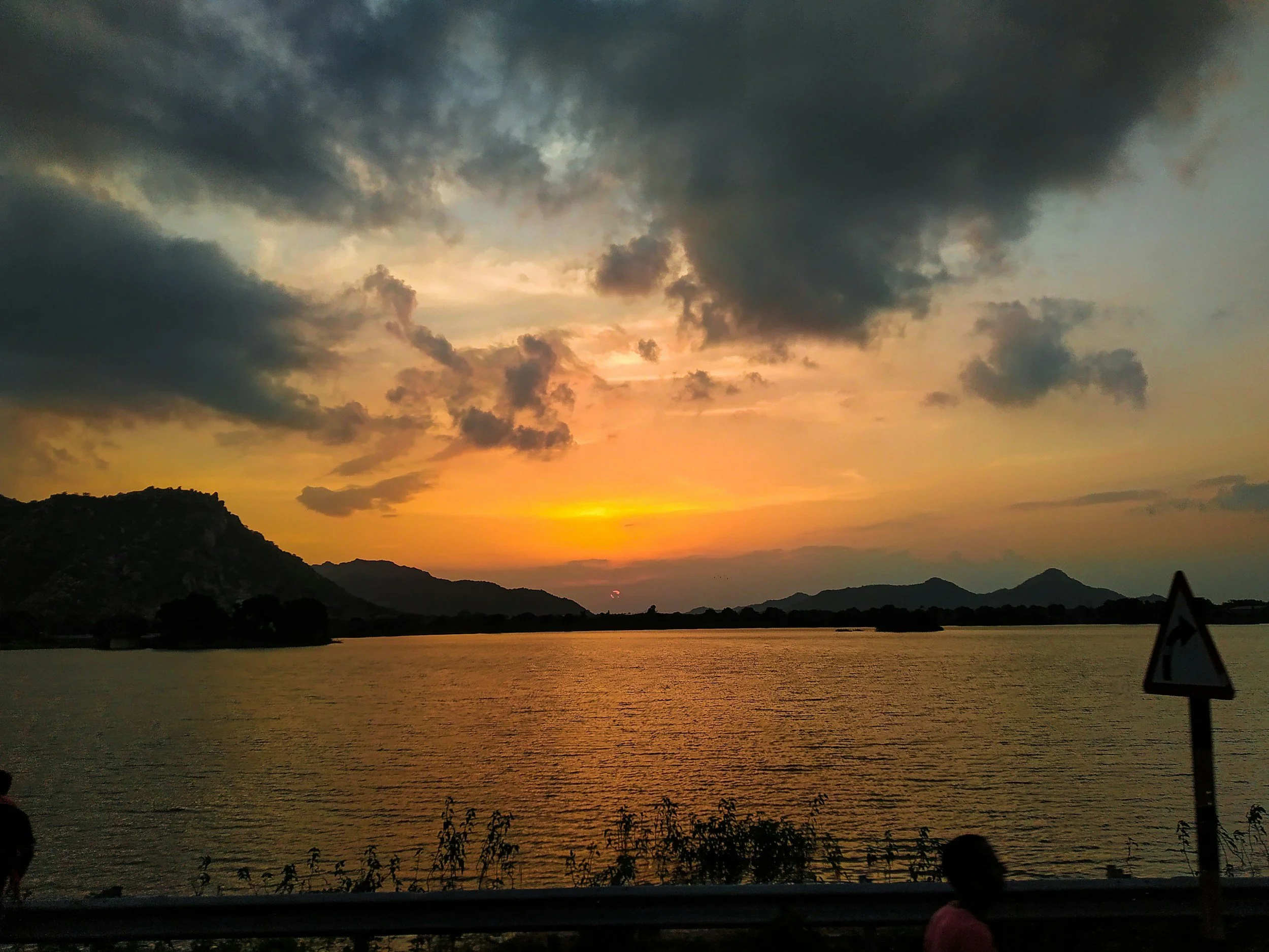 A sunset over a body of water with mountains in the background. Dark clouds are scattered across the sky, and the water reflects the sunset colors. There is a road with a traffic sign on the right side and a person standing near the bottom left.