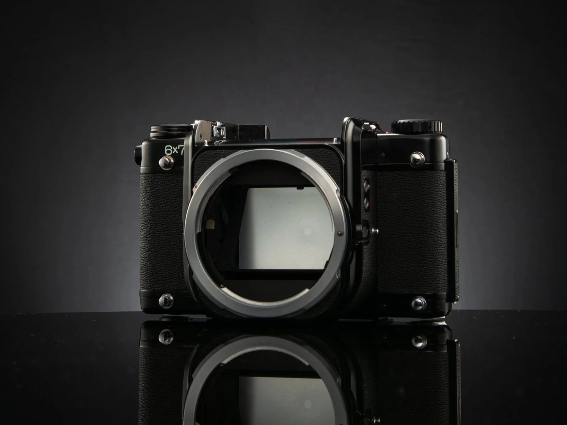 A black digital camera body without a lens, placed on a reflective surface with a dark, gradient background.