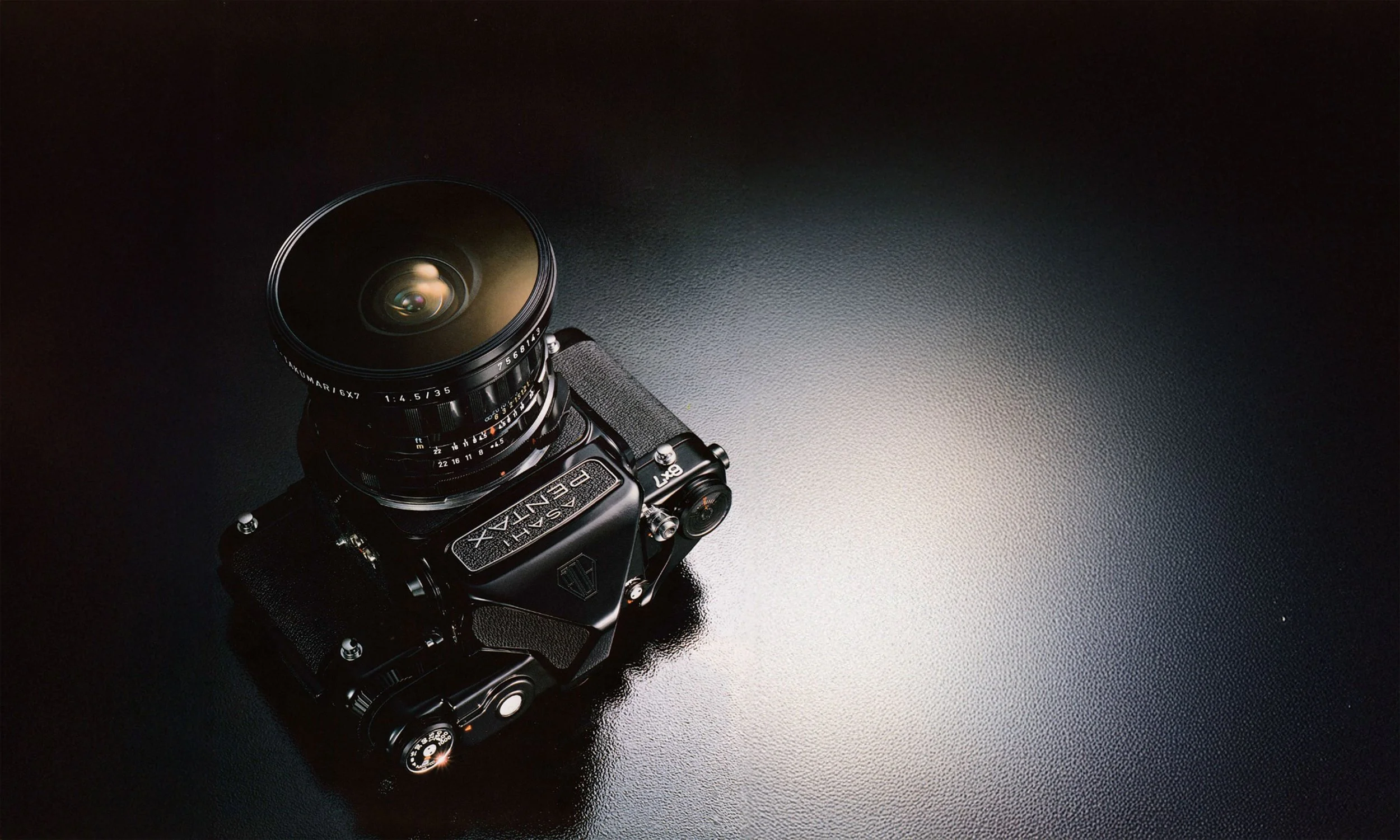 A black digital camera with a large lens, resting on a reflective black surface with a textured finish.